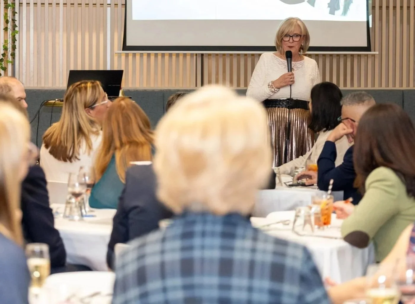 Kerryn Powell speaking into a microphone at a presentation or conference. Audience members seated at round tables are listening to her, with some taking notes or enjoying drinks.