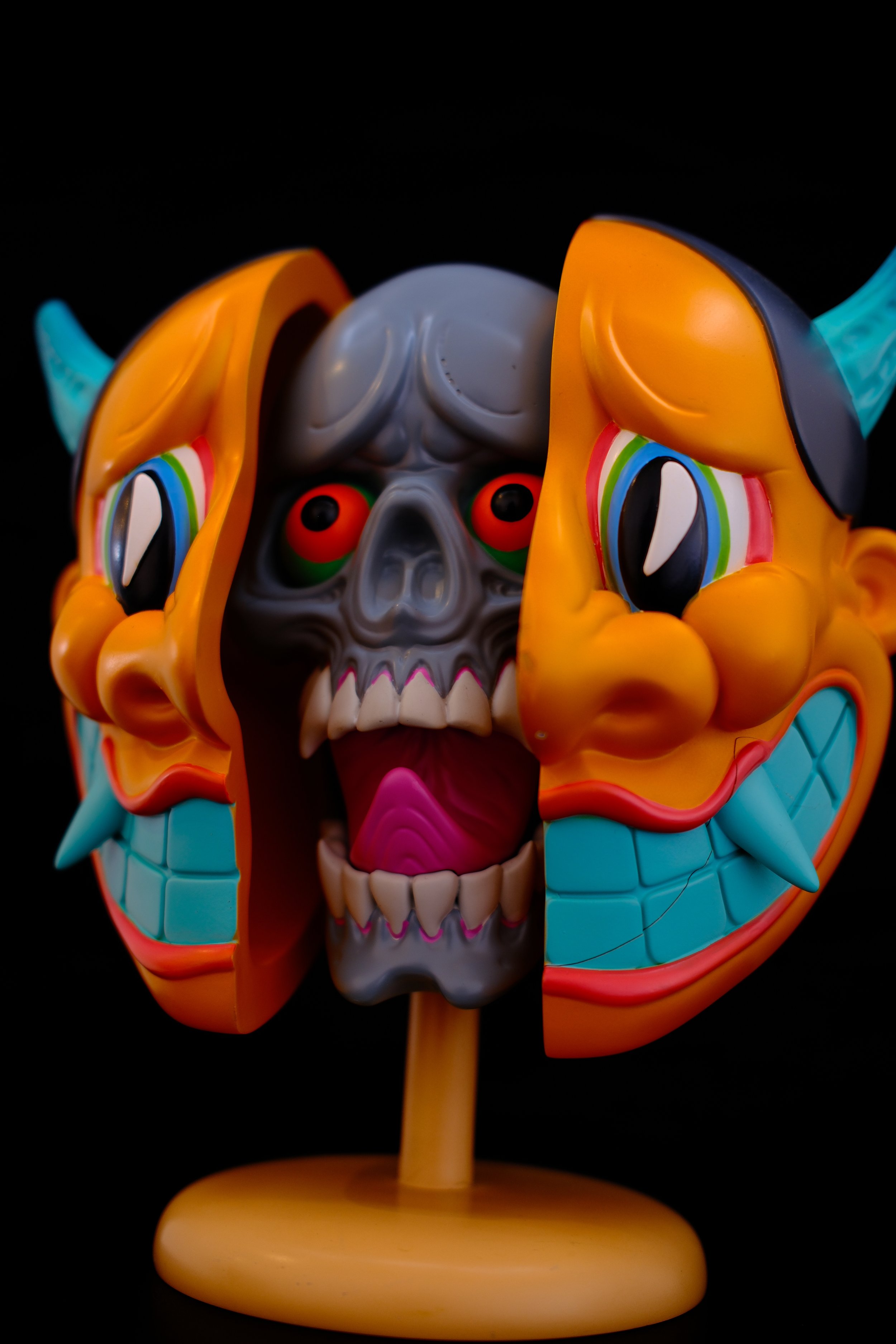 Colorful, spooky skull mask with clown-like features, bright orange cheeks, large blue teeth, and rainbow eyes, mounted on a wooden stand against a black background.