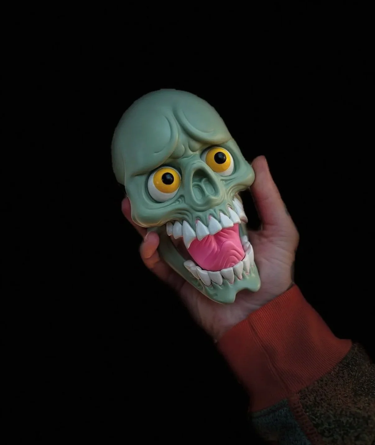 Hand holding a green zombie skull mask with large yellow eyes, sharp teeth, and a pink tongue against a black background.