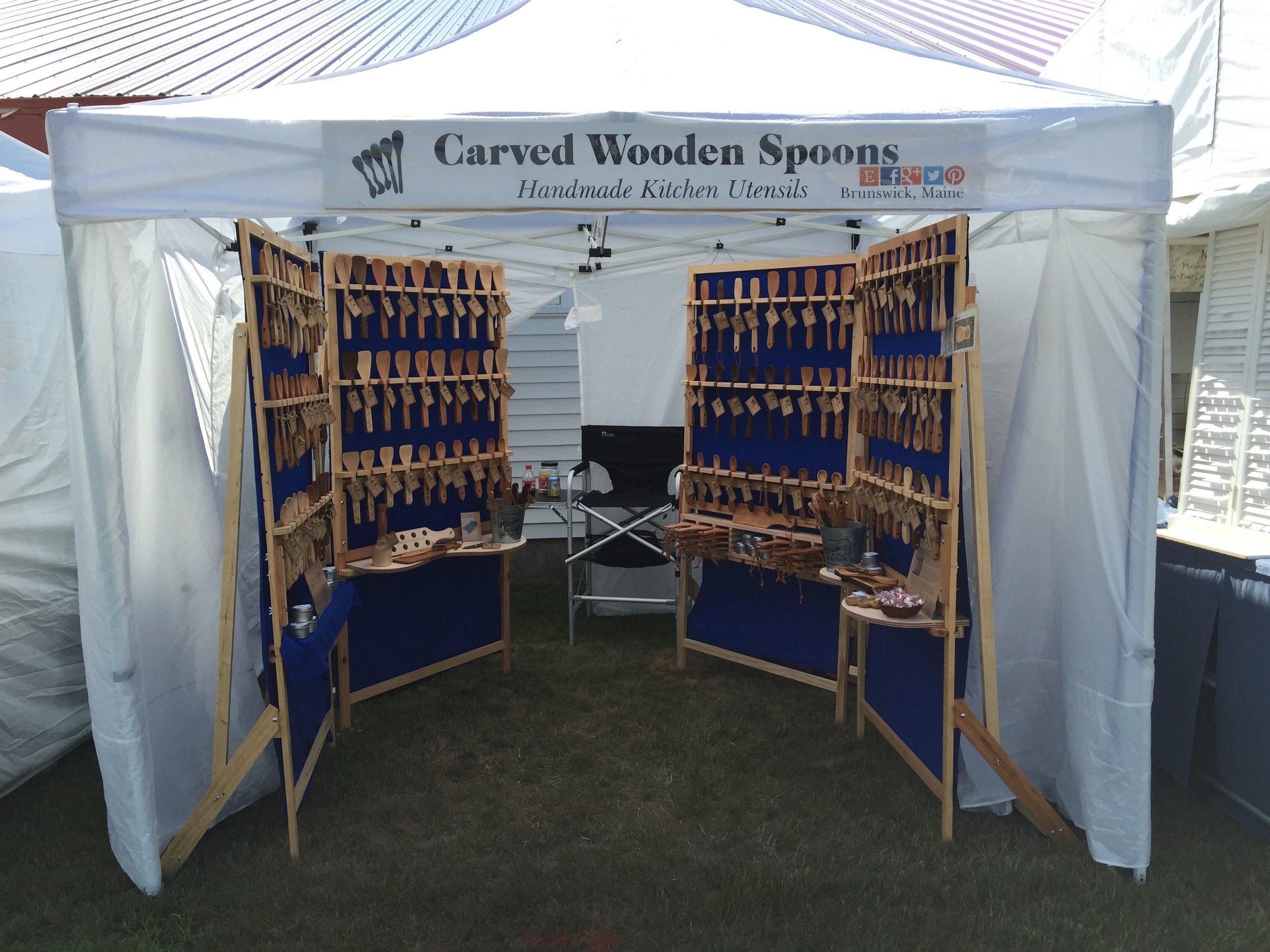 A display booth selling carved wooden spoons, with multiple shelves filled with wooden utensils under a white canopy at an outdoor event.