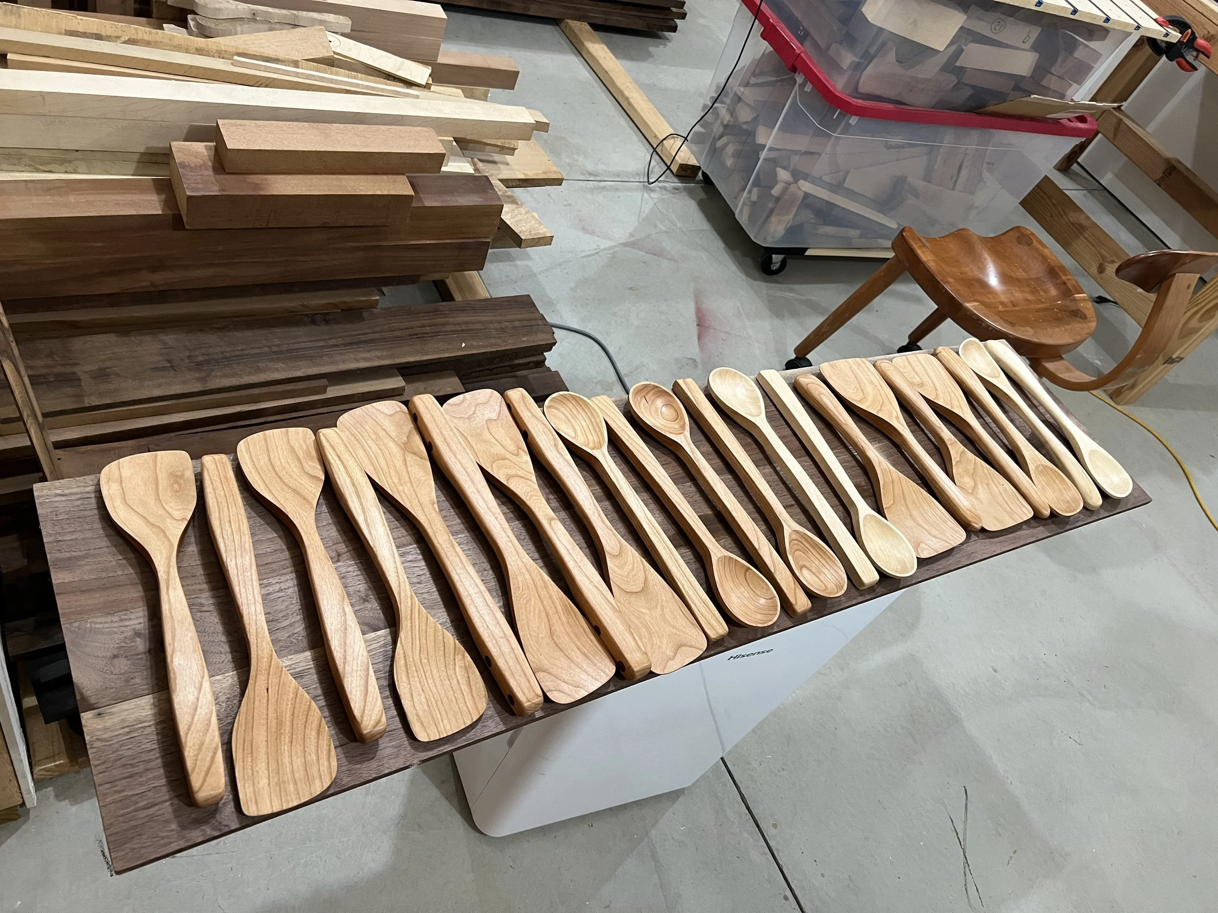 A row of wooden spoons and spatulas arranged on a dark wood table in a woodworking shop, with various wooden pieces and tools in the background.