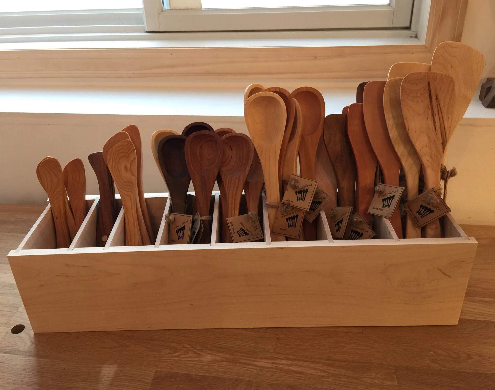 A wooden display box containing various wooden spoons arranged vertically by size and color.