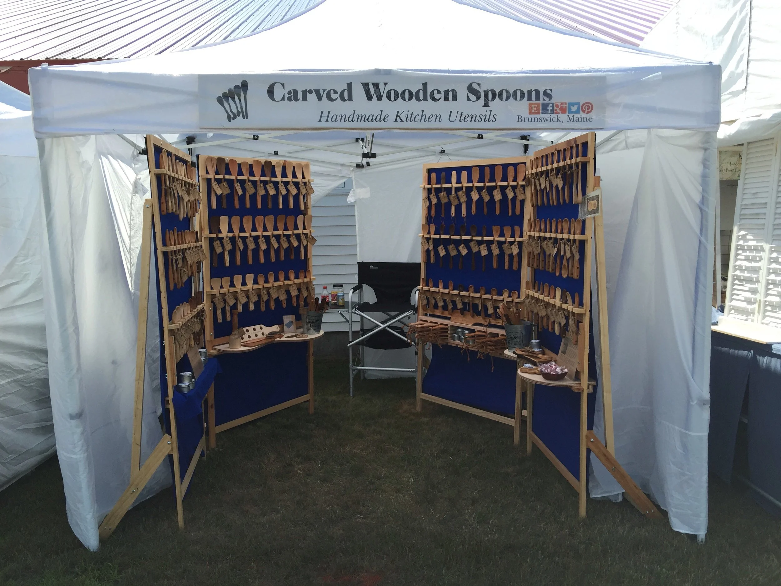 A booth selling handmade carved wooden spoons at an outdoor market. The booth has a white canopy with a sign that reads 'Carved Wooden Spoons, Handmade Kitchen Utensils' from Brunswick, Maine. Wooden spoons are displayed on blue backdrops inside wooden frames.