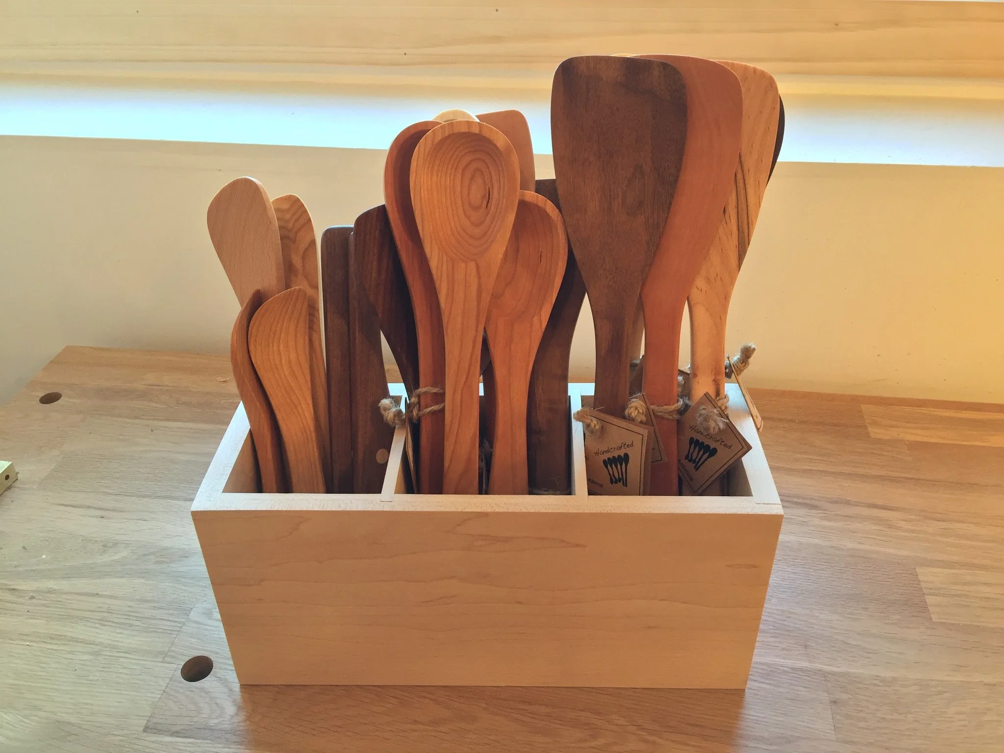 A wooden box containing various sizes of handcrafted wooden spoons on a wooden table.
