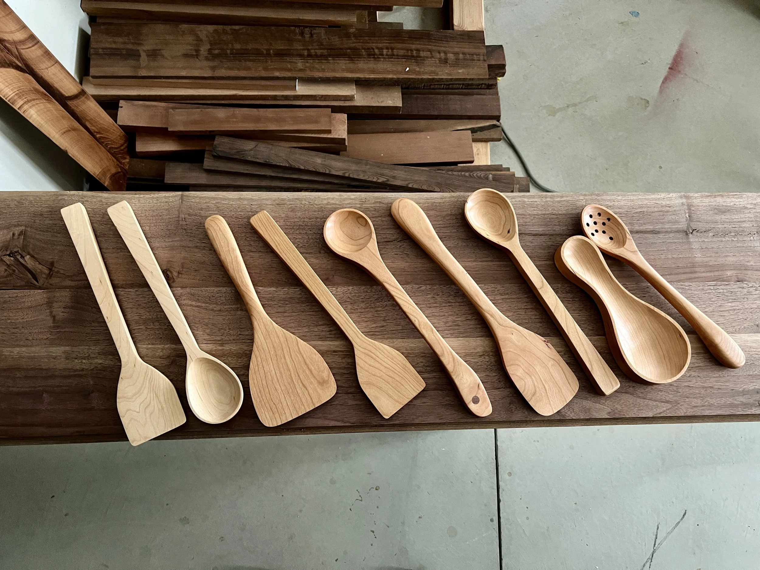 Nine wooden kitchen utensils laid out on a wooden work surface, including spatulas, scoops, and spoons, with some having differing shapes.