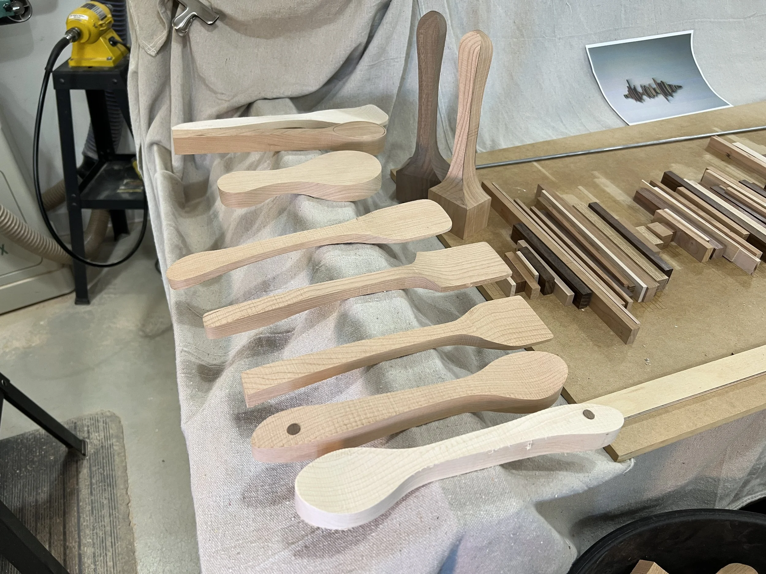 Various unfinished wooden spatulas and paddles laid out on a fabric-covered work surface, with woodworking tools and small wooden pieces in the background.