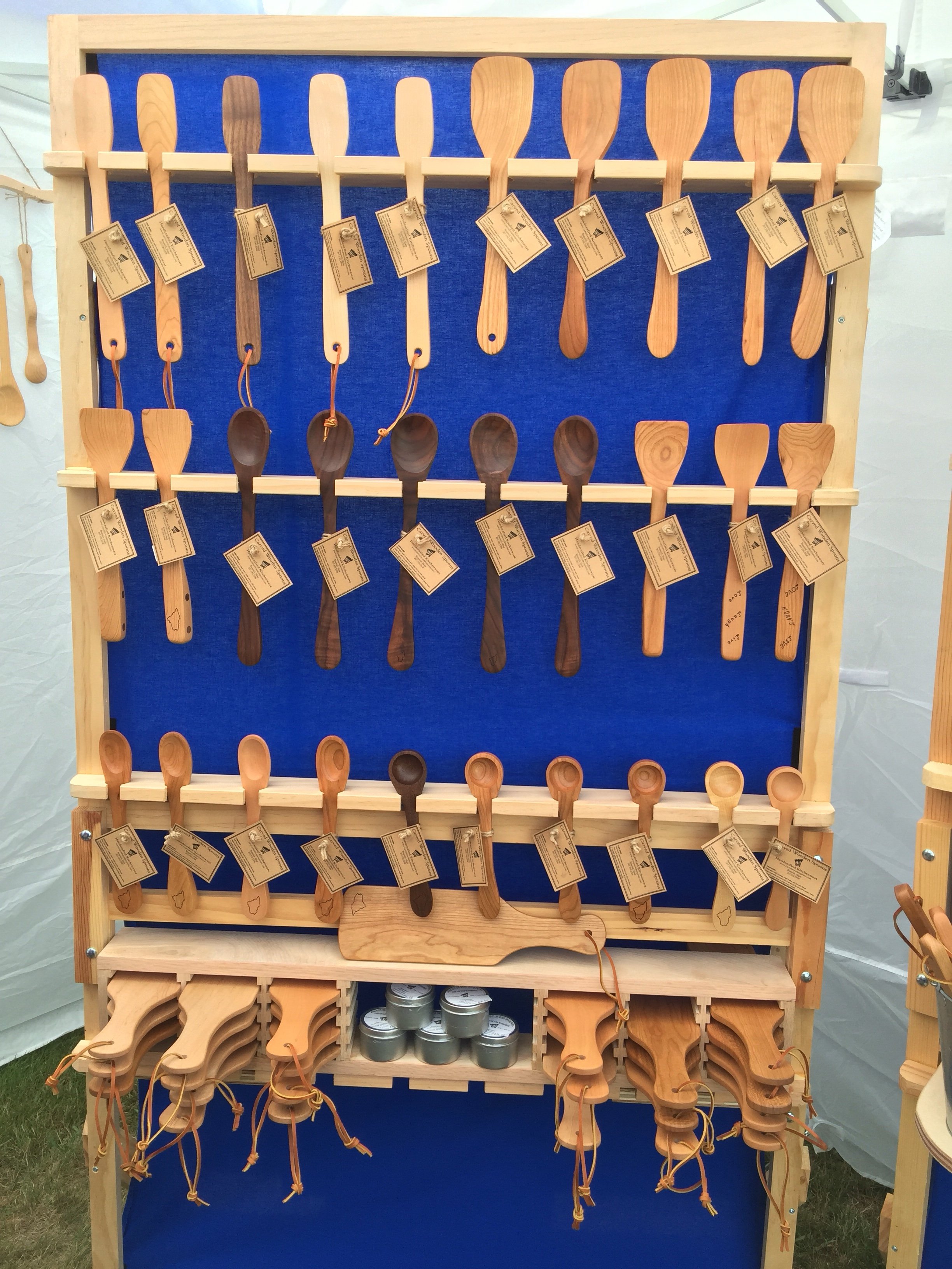 Wooden spoons and spatulas on display at a craft fair with a blue backdrop and price tags attached.