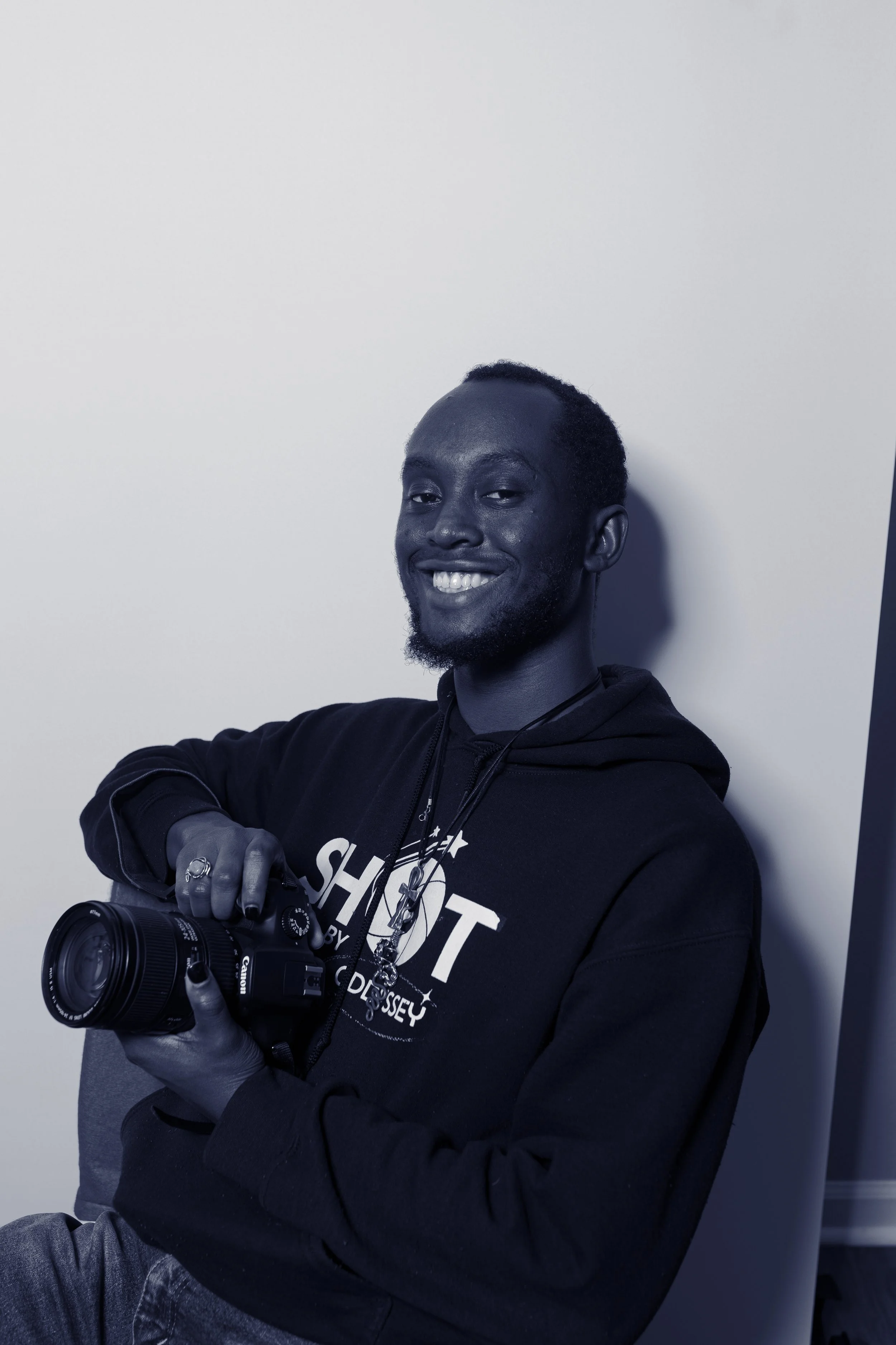 A smiling man sitting against a white wall, holding a Canon camera. He is wearing a black hoodie with white text and appears to be in a casual setting.