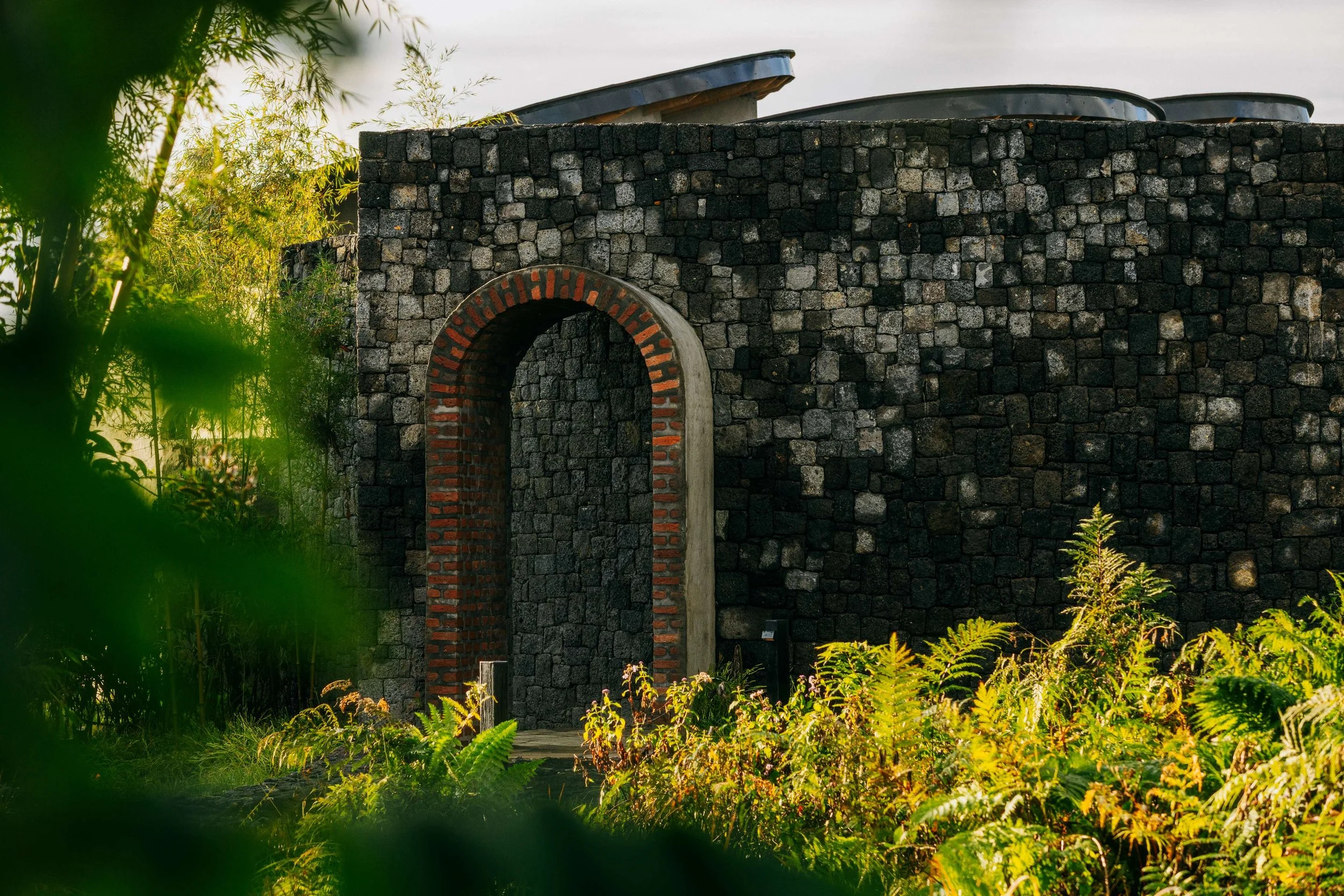Bisate Sanctuary's arched brick entrance set into a volcanic stone wall, framed by lush ferns and bamboo at Volcanoes National Park, Rwanda — Black Sable