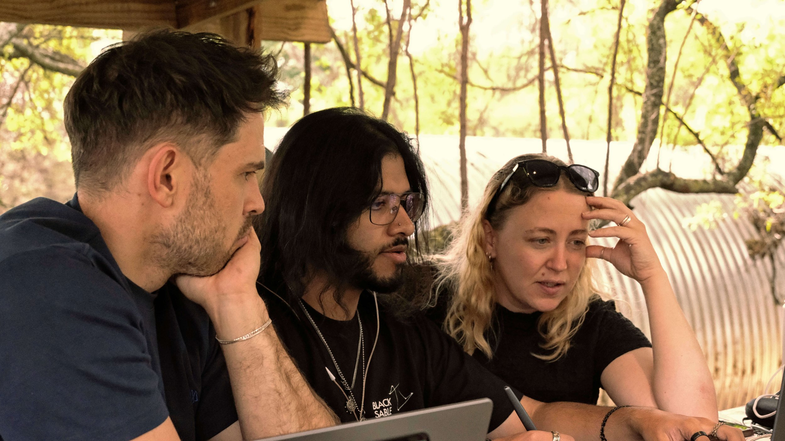 Three Black Sable team members in focused conversation around a laptop in a covered outdoor studio space, surrounded by natural bushveld.