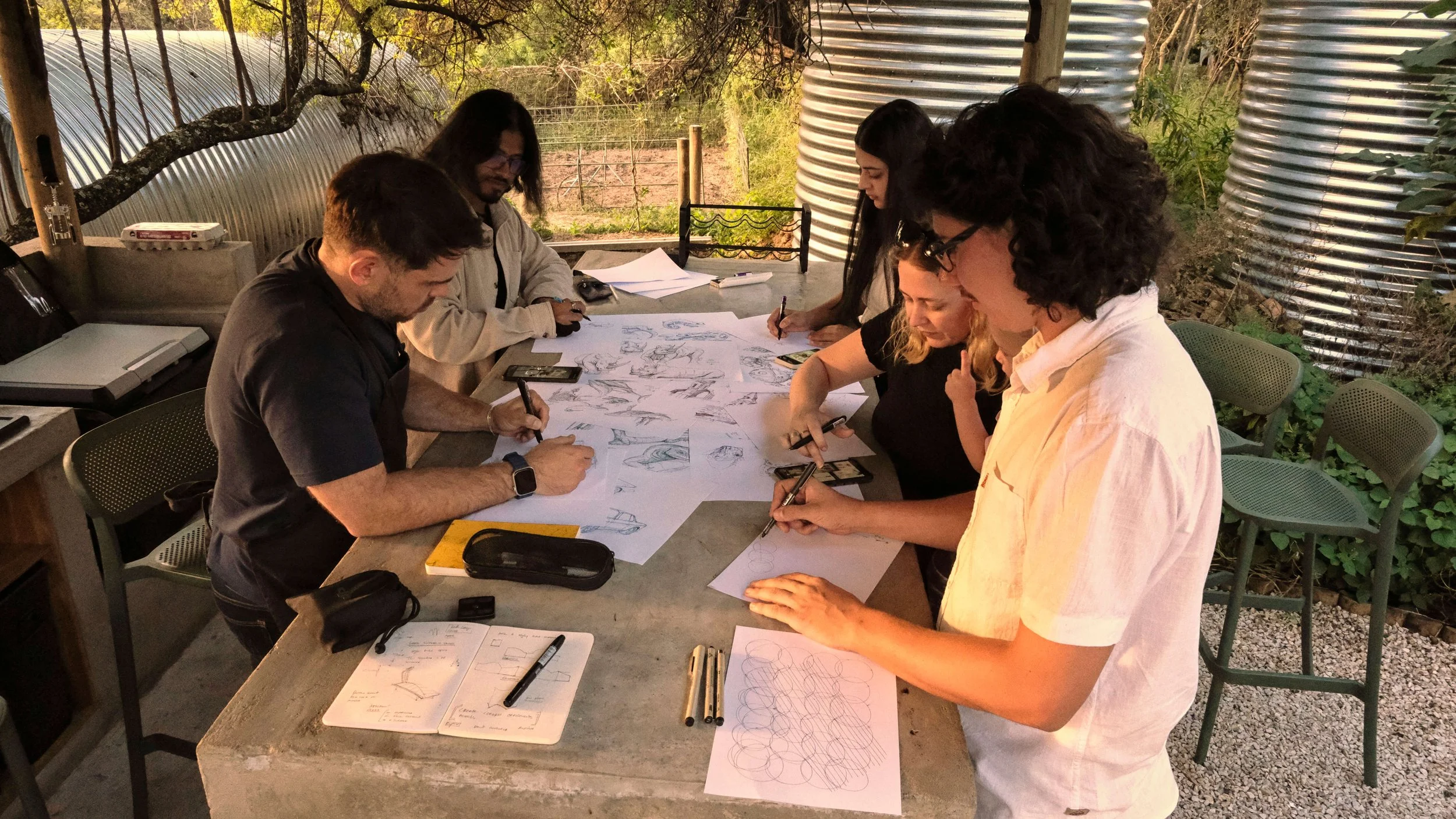 Five Black Sable team members seated around a concrete table in an outdoor studio, all drawing simultaneously on large sheets of paper spread across the surface.