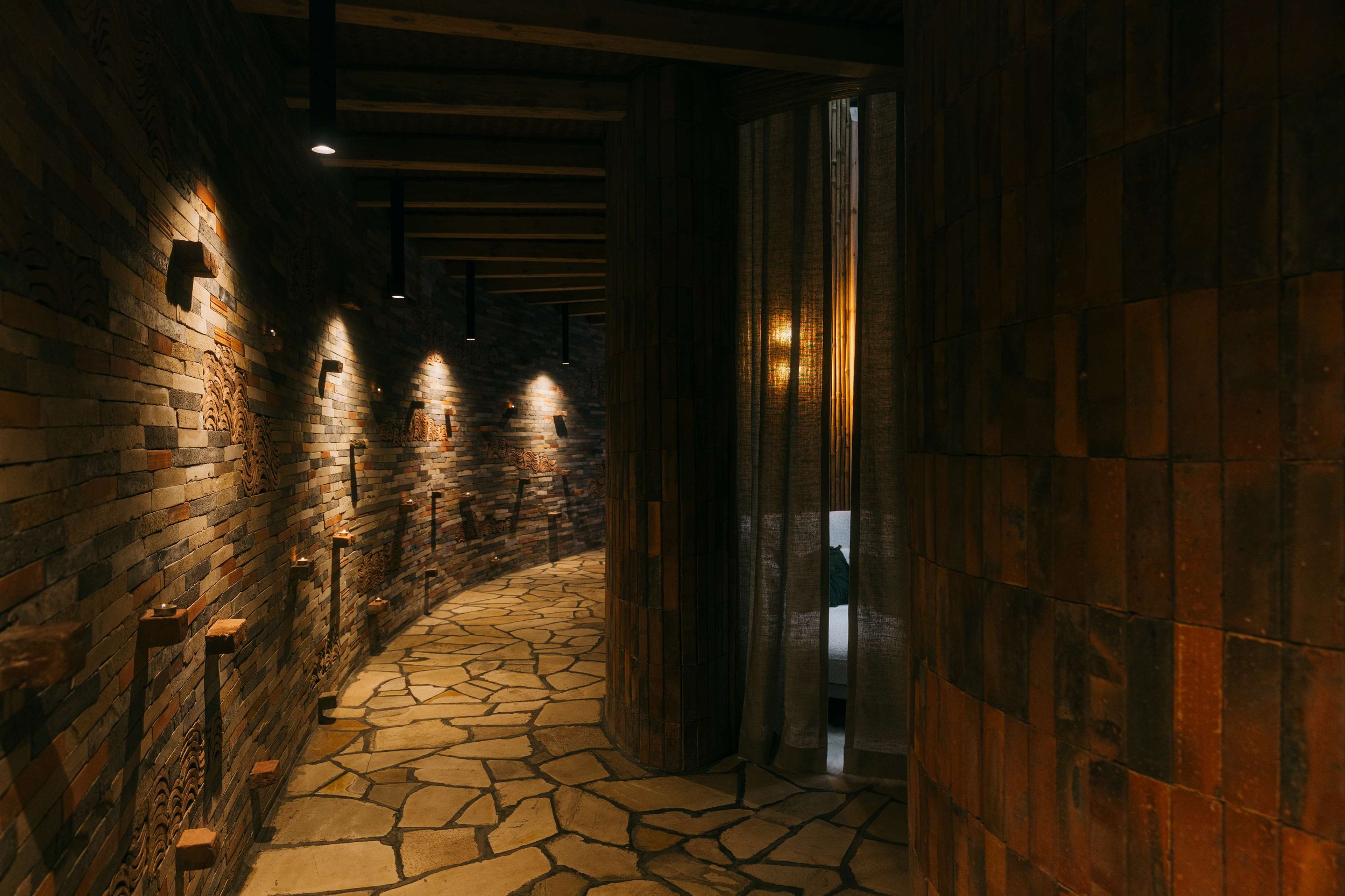 Dark, textured corridor inside Bisate Sanctuary's Spa wing, lined with handcrafted brick walls, carved stone details, candlelit sconces and a flagstone floor — Black Sable