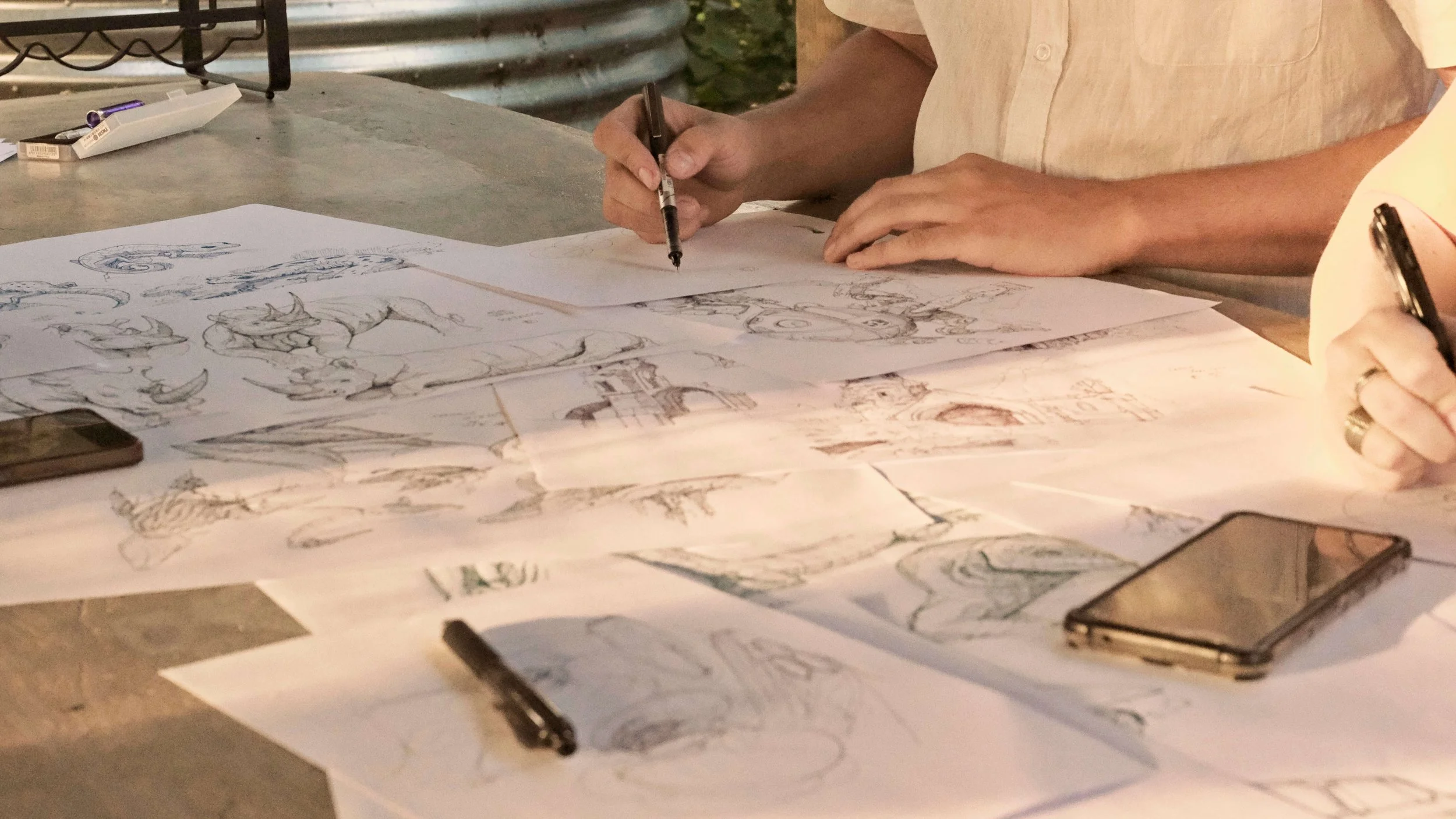 Close-up of a designer's hands working on detailed pen-and-ink architectural and wildlife sketches spread across a table, with phones and drawing tools nearby.
