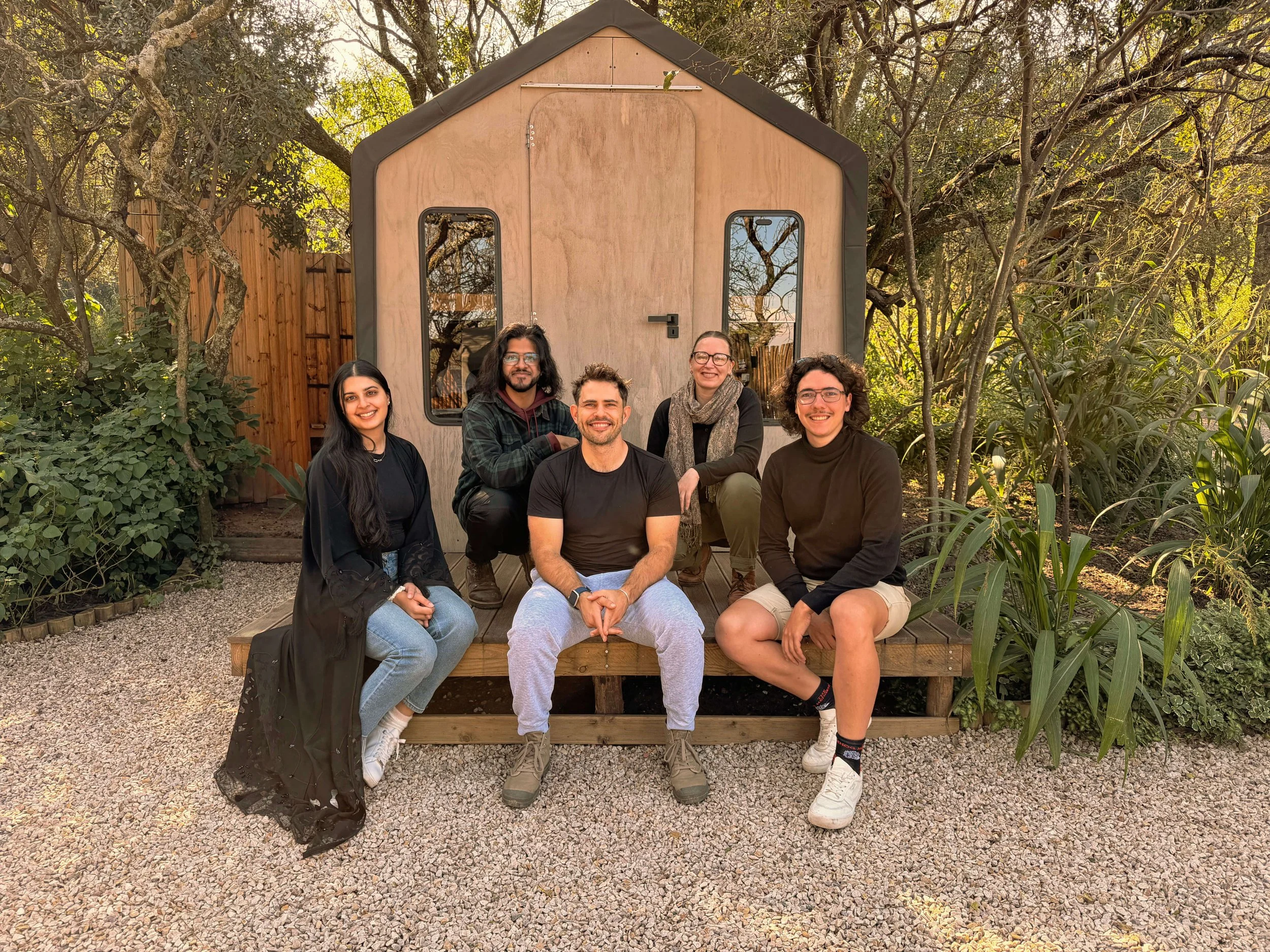 Five members of the Black Sable team sitting and standing in front of a small timber studio structure surrounded by trees and gravel landscaping.