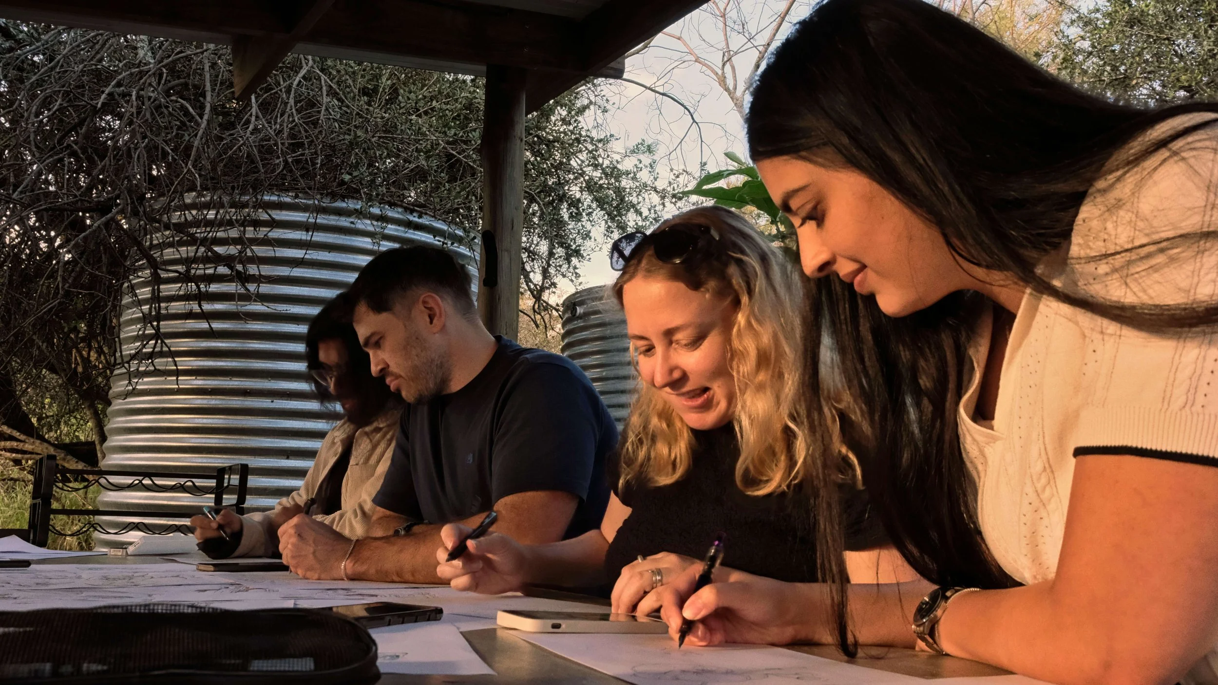 Four Black Sable team members seated side by side in an open-air studio, each drawing quietly at a shared table, with corrugated water tanks and bushveld visible behind them.