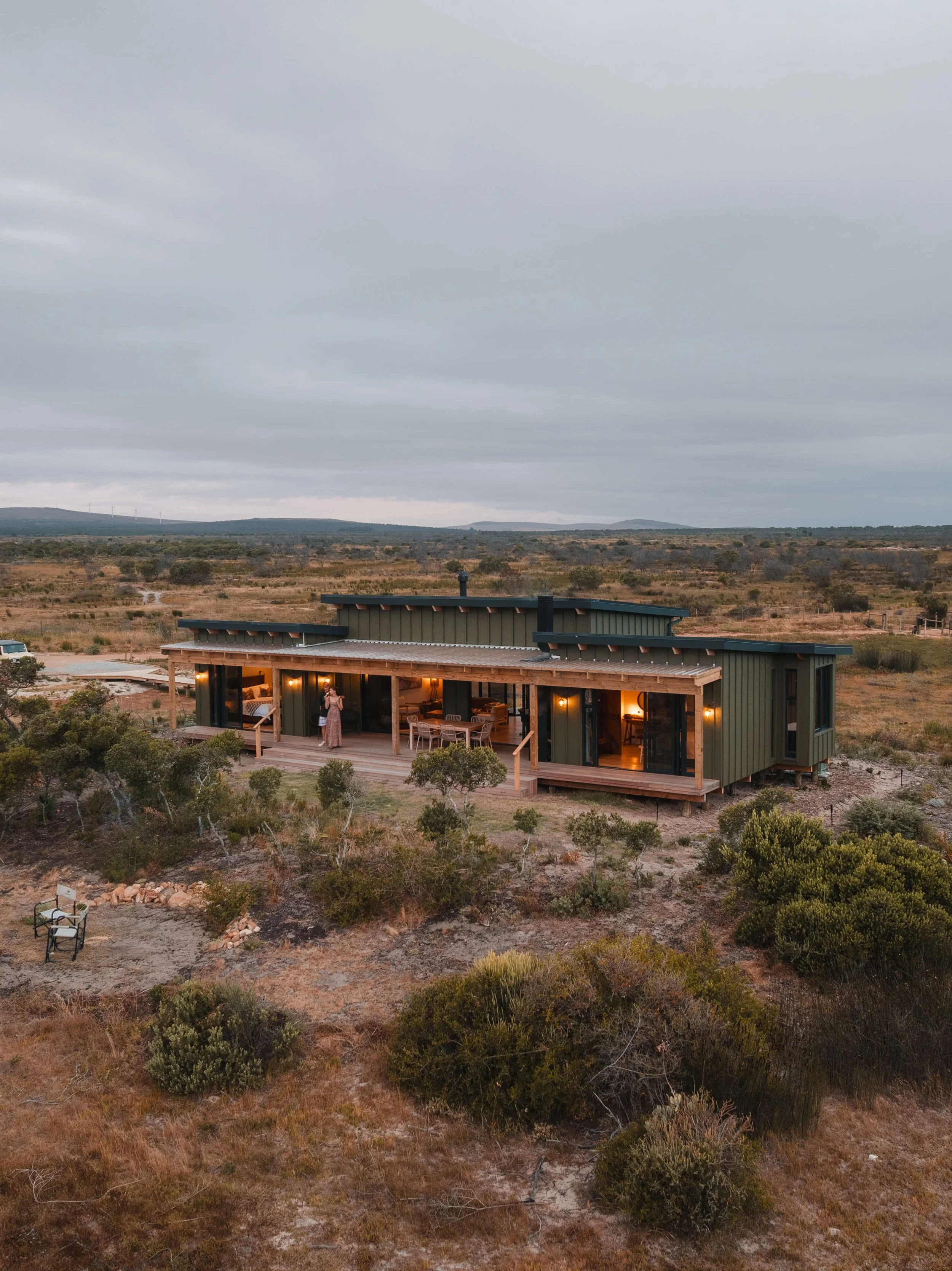 Elevated exterior view of The Salt Mine at dusk, showing the full timber-decked veranda, dark cladding and warm interior lighting against the open fynbos landscape