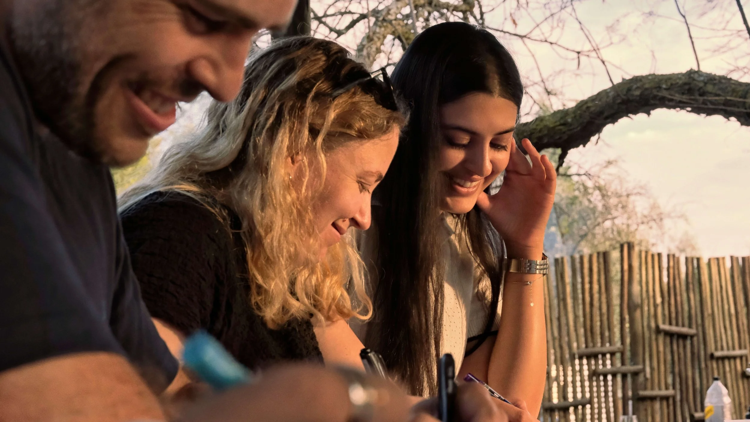 Three Black Sable team members laughing together while sketching outdoors, bathed in warm golden light with a wooden fence and bare tree branches in the background.