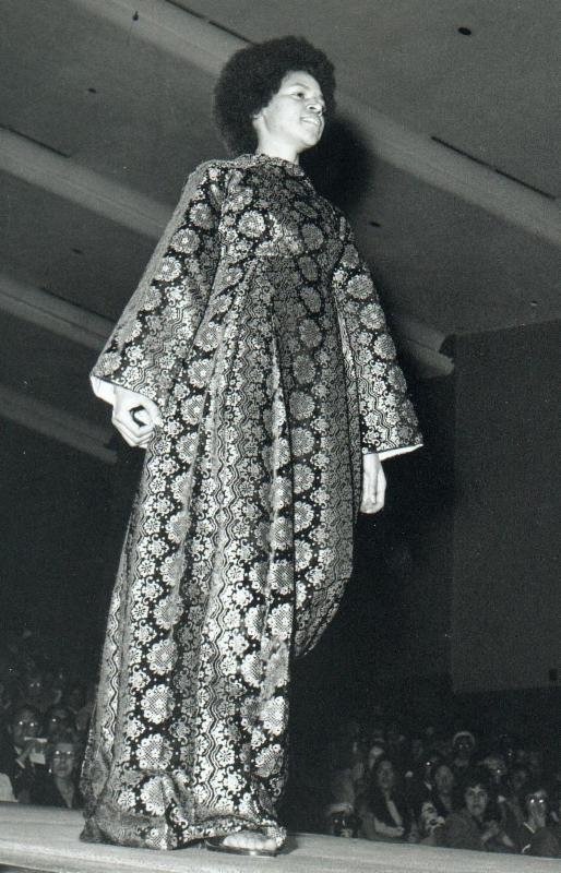 Model walking down runway in floor length, long sleeve, patterned dress 