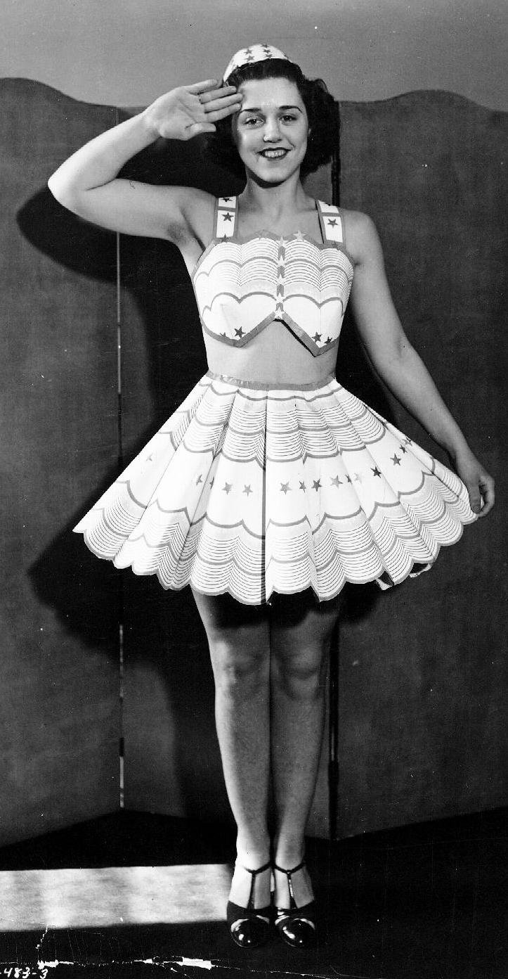 Model wearing knee-length skirt and crop top with hat, saluting 