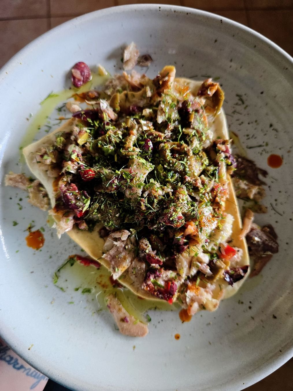 Overhead view of sesame crackers topped with sardines, muffaletta olive salad, dried herbs, and olive oil on a ceramic plate