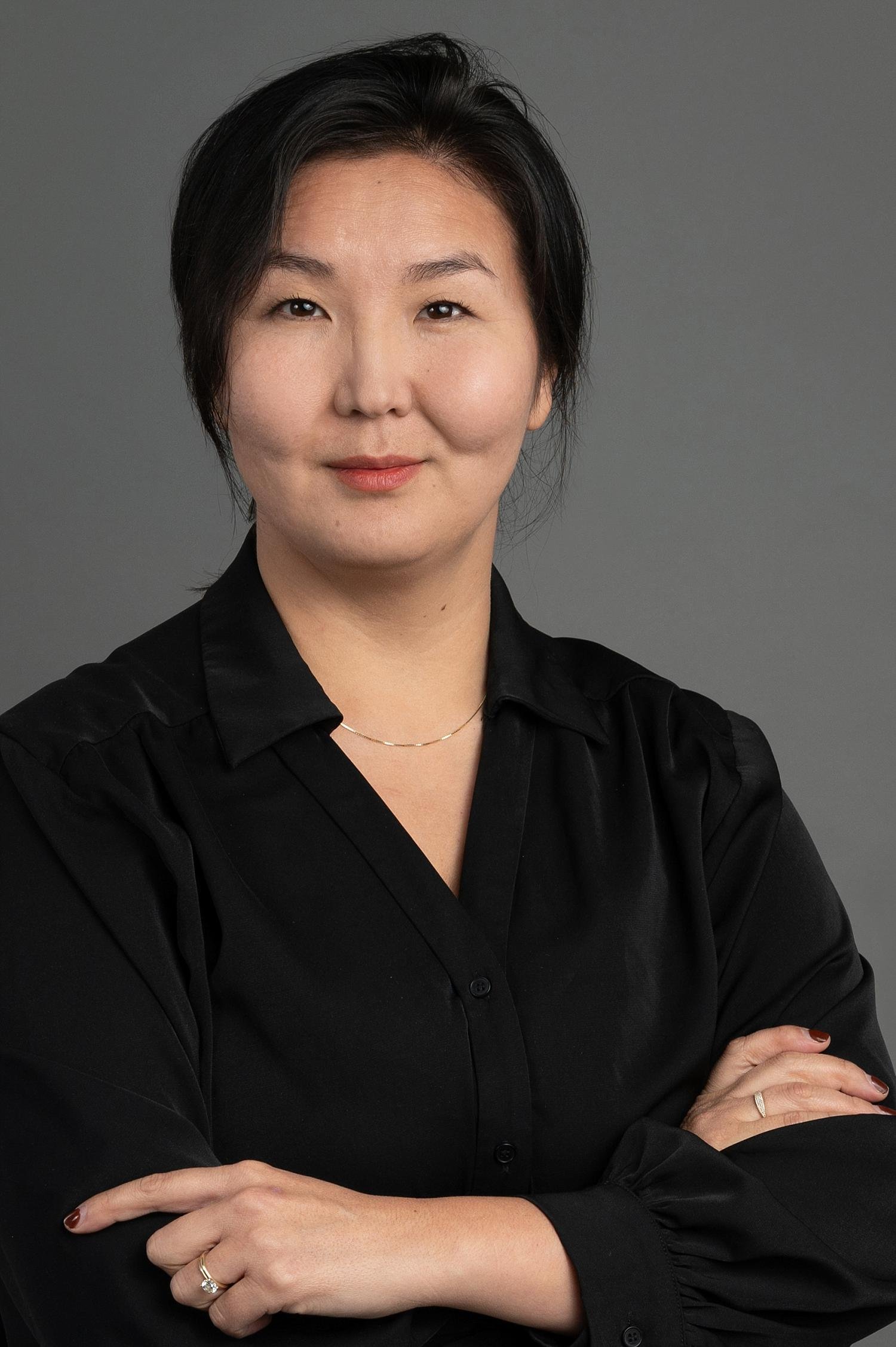 A woman with short black hair and light skin, wearing a black blouse and a delicate necklace, standing against a plain gray background.