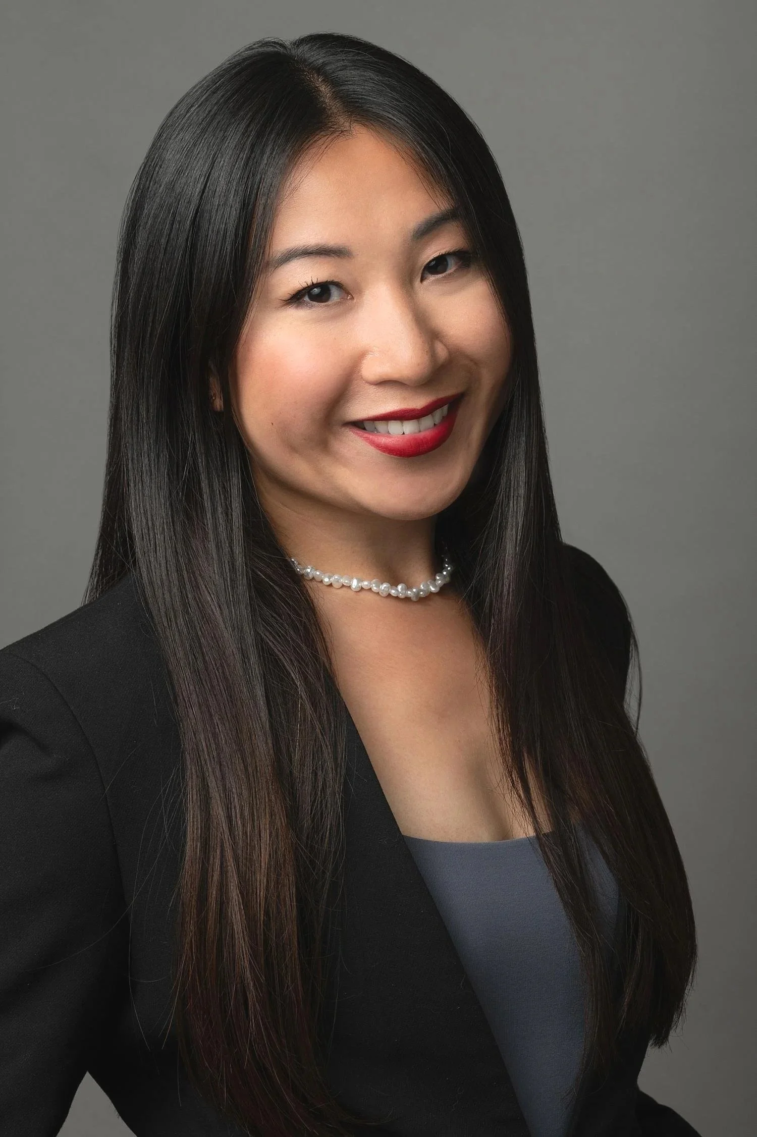 Headshot of an Asian woman with long black hair, wearing a black blazer, pearl necklace, and gray top, smiling against a plain gray background.