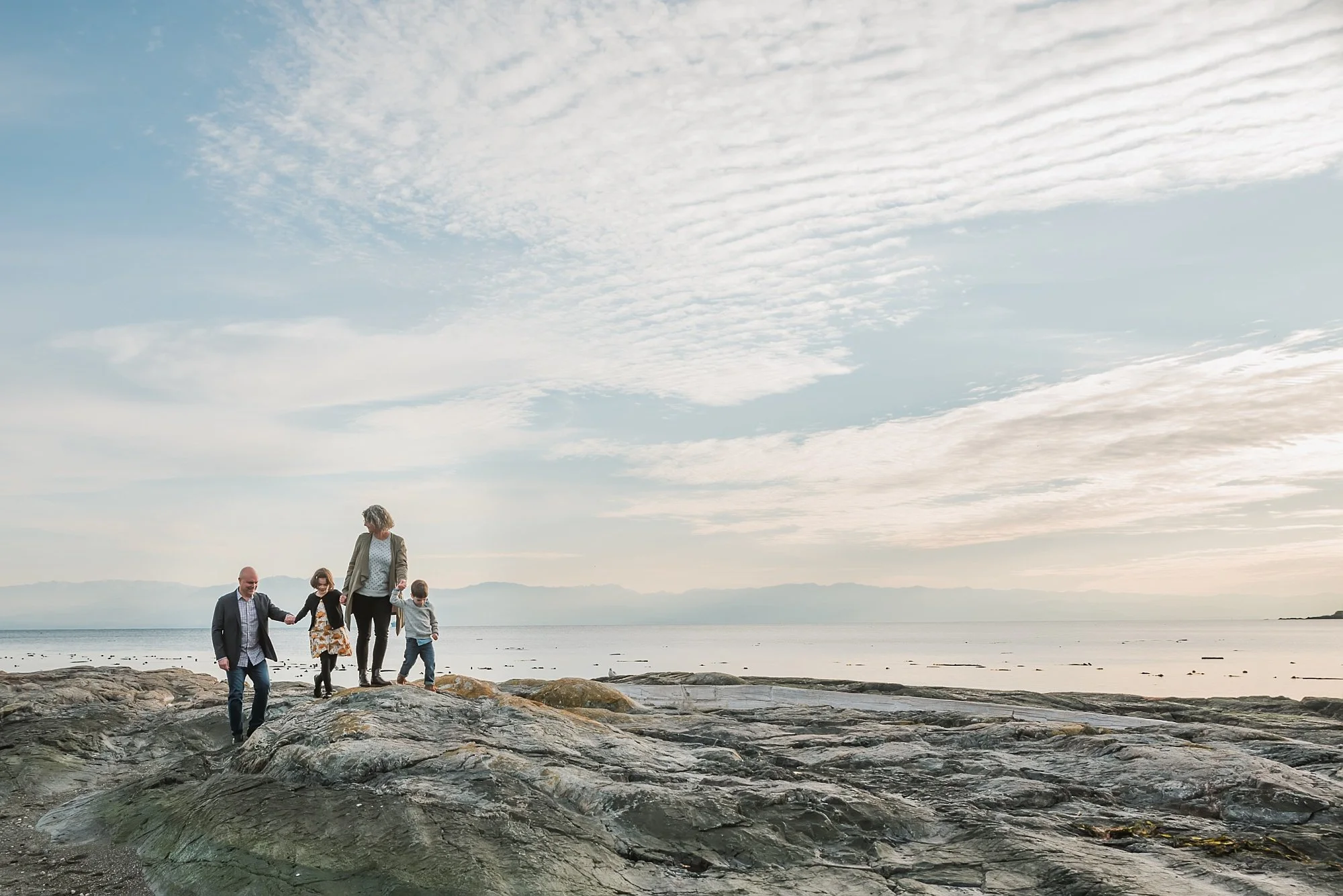 Family walking on rocky beach near ocean under cloudy sky.
