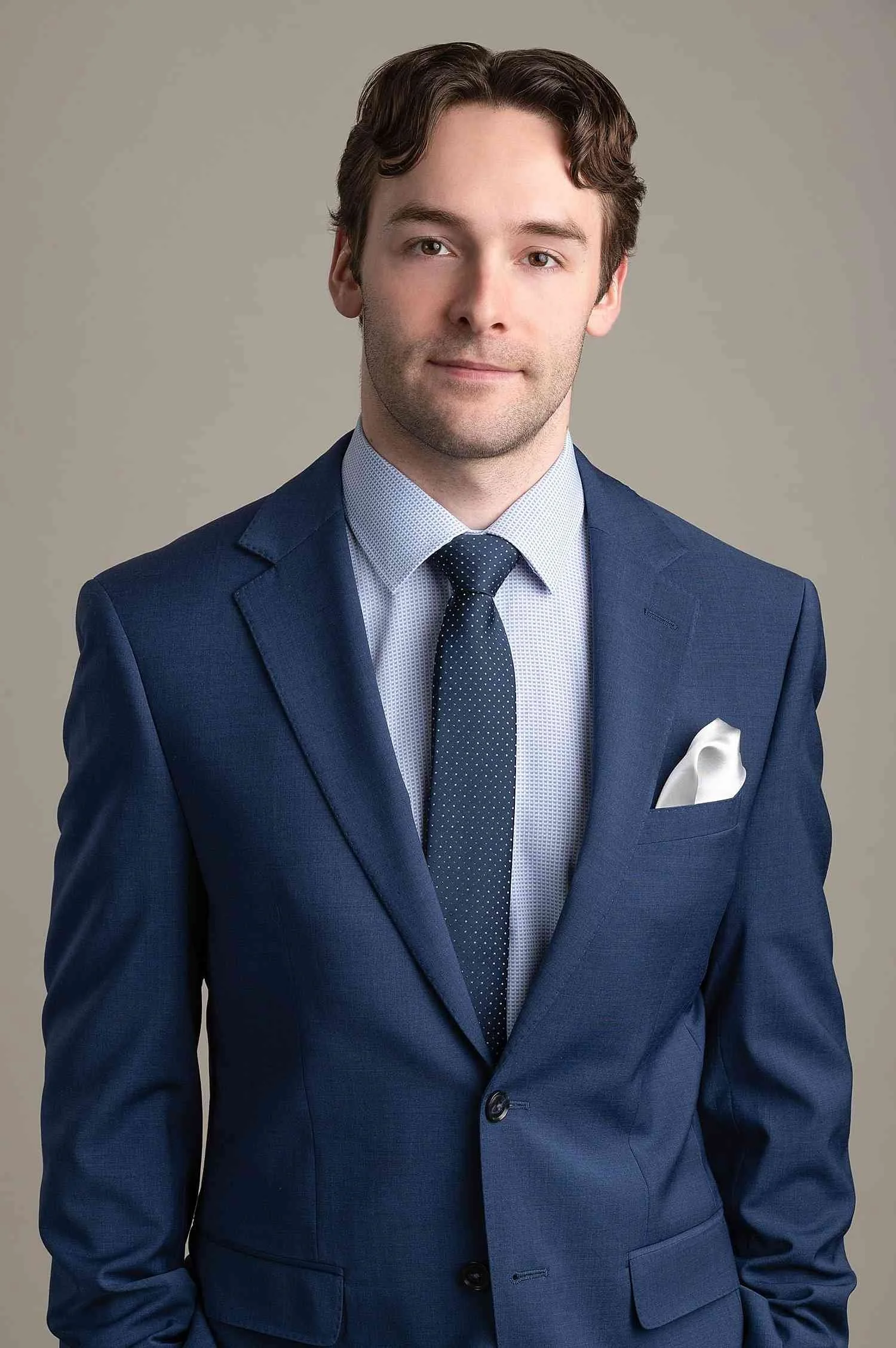 A young man with brown hair wearing a navy blue suit, a light blue patterned dress shirt, a dark blue dotted tie, and a white pocket square, standing against a beige background.
