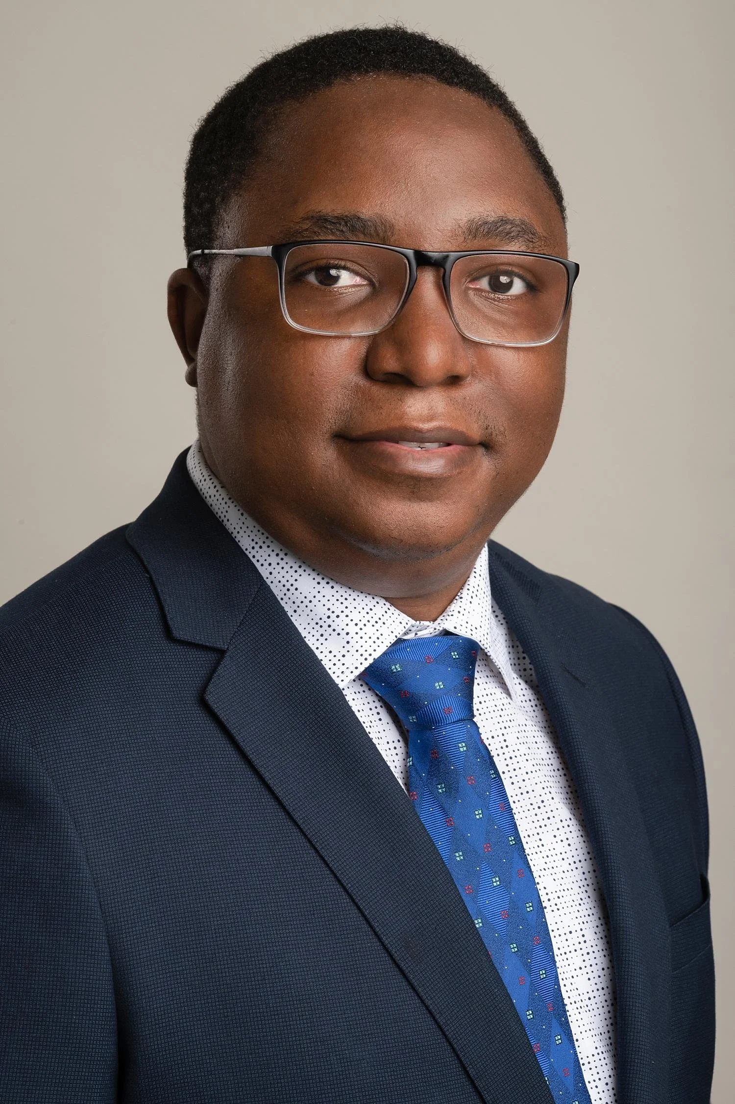 A professional headshot of a man wearing glasses, a dark suit, a white shirt with black dots, and a blue tie with red and white patterns.