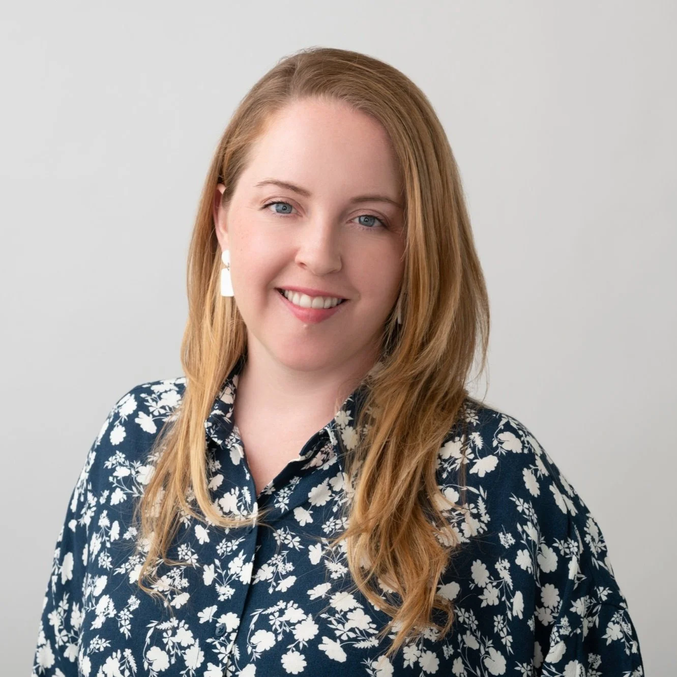 Person with long blonde hair wearing a floral-patterned shirt and white earrings, smiling against a plain background.