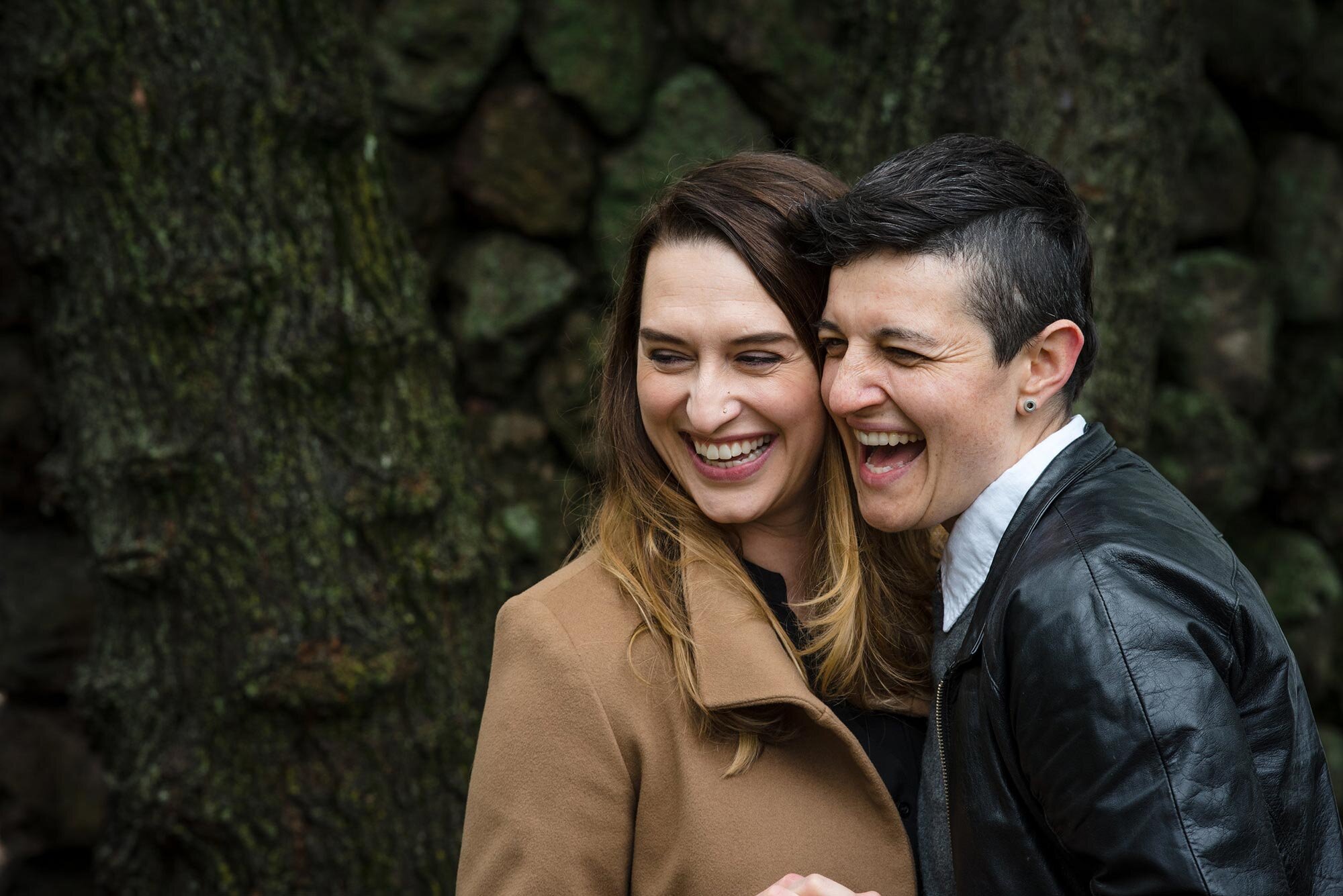 Couple laughs together in front of stone wall during autumn queer family photo session Michelle Schapiro Photography