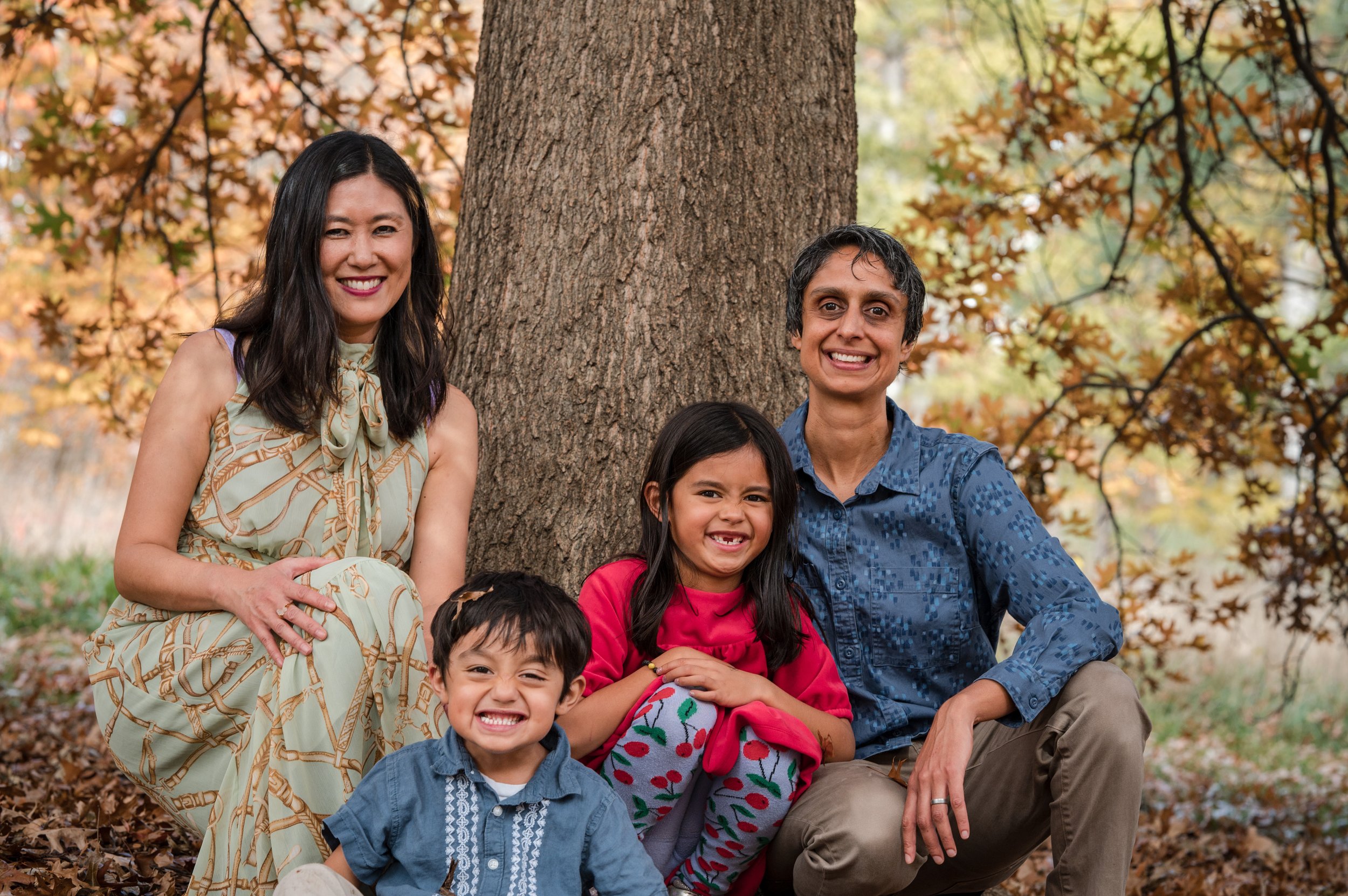 LGBTQIA+ family featuring two smiling kids and two happy parents sitting at the base of a tree in boston park during the fall by Michelle Schapiro Photography