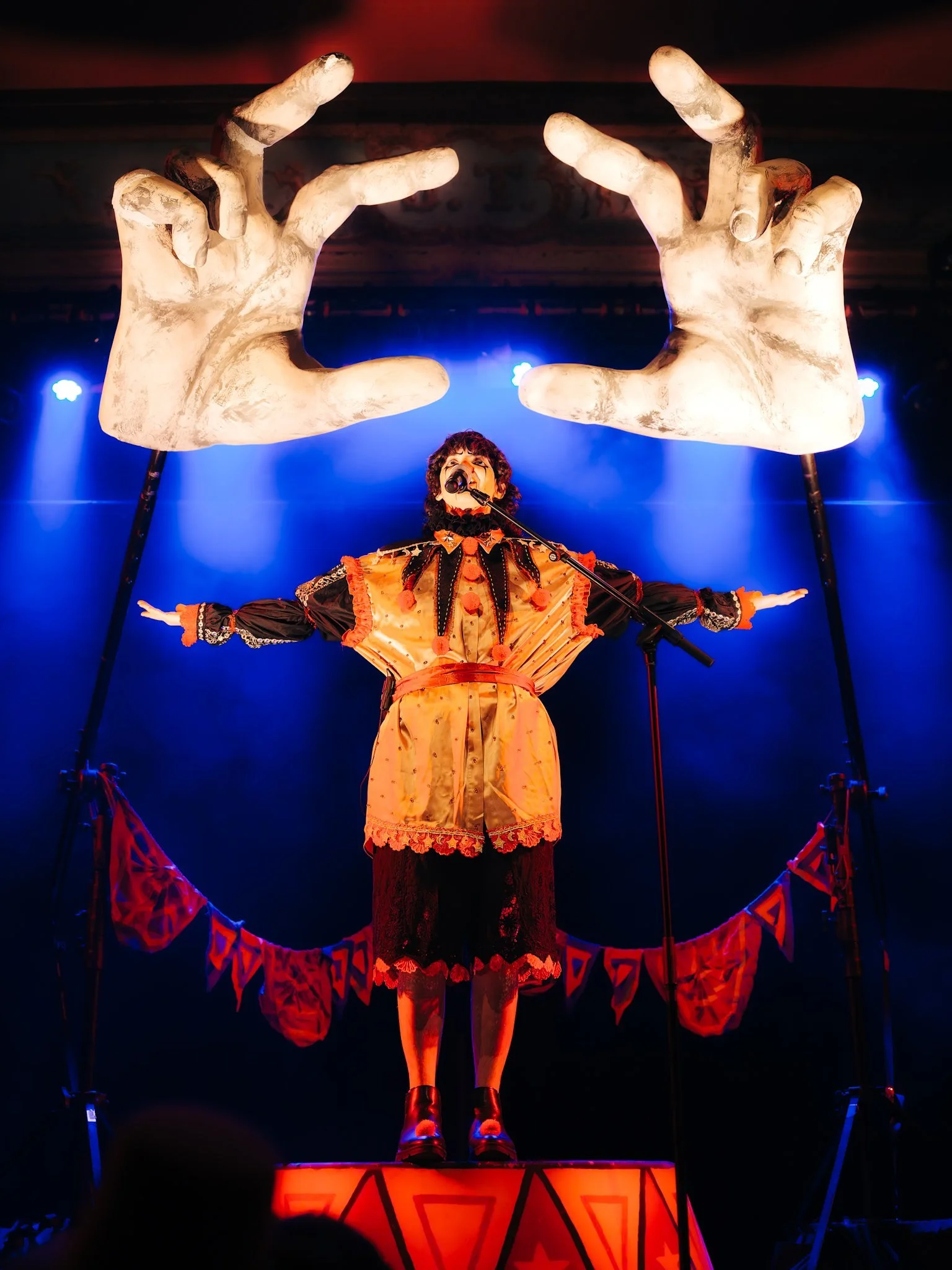 Madilyn Mei in a clown costume with arms raised under giant cartoon hand props on stage. Performing at Bluebird Theater in Denver, CO
