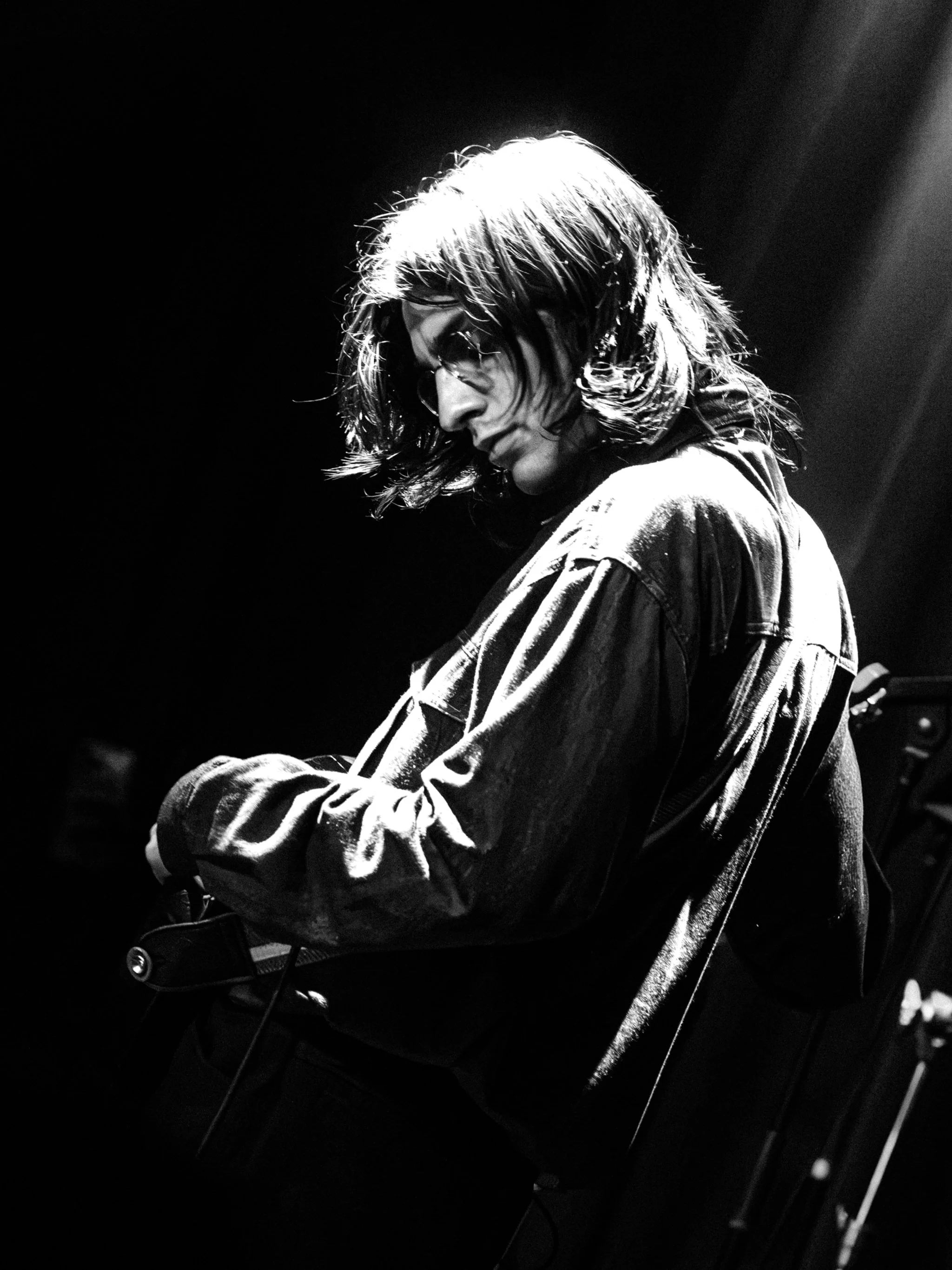 Black and white photo of a long-haired guitarist backlit on a dark stage, head down