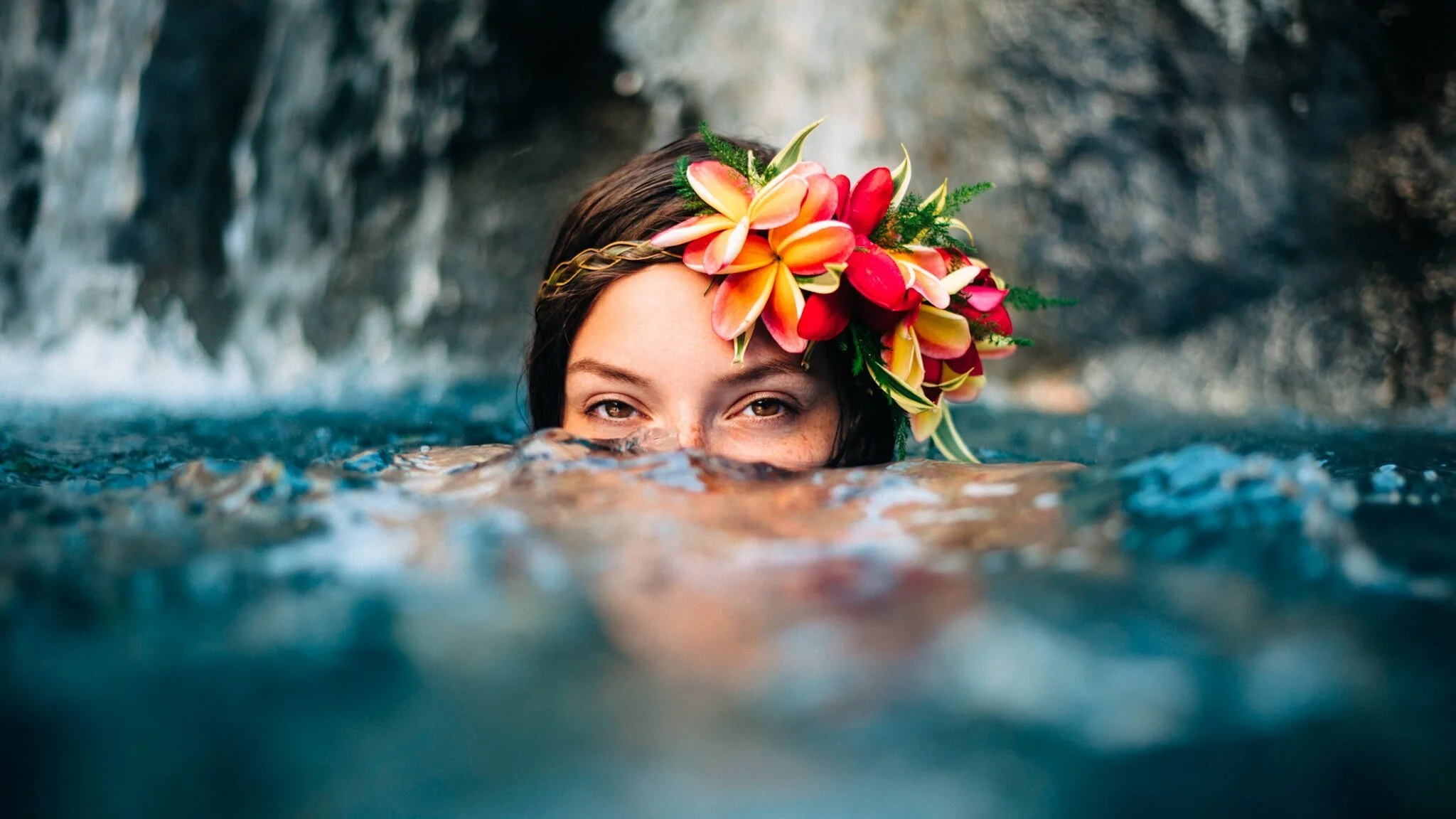 Woman with multi-colored flowers in her hair half-submerged in water in front of a waterfall