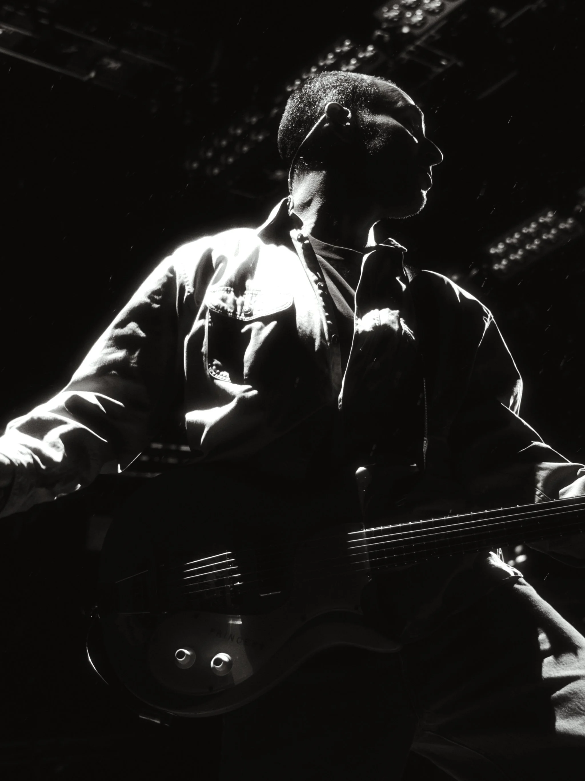Black and white photo of a guitarist rim-lit from behind, bass slung across his chest