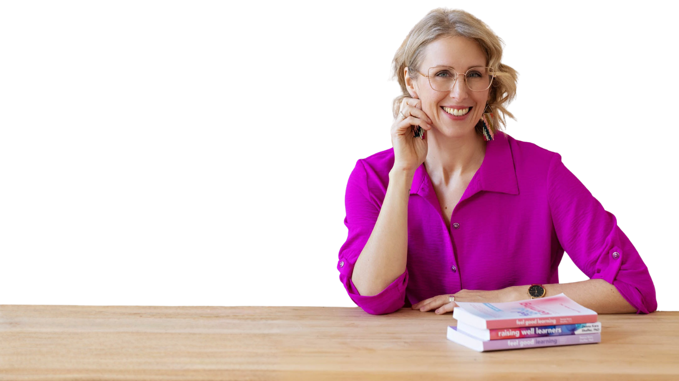 A woman with blonde hair and glasses smiling, sitting at a wooden table with three books stacked in front of her, wearing a vibrant pink shirt.