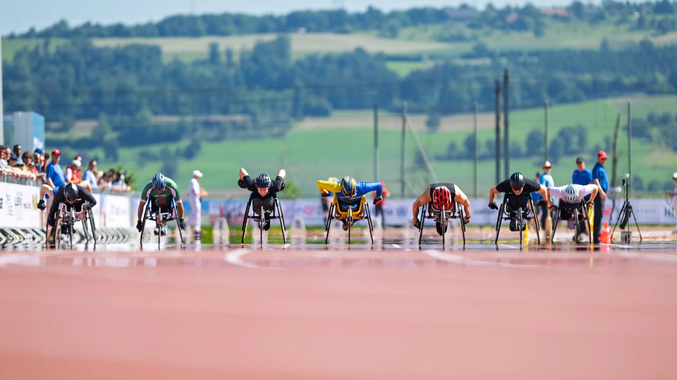 Eight T52  wheelchair athletes race down the track in a 100m event. It is a tight race to the line. There is a green hill in the background.