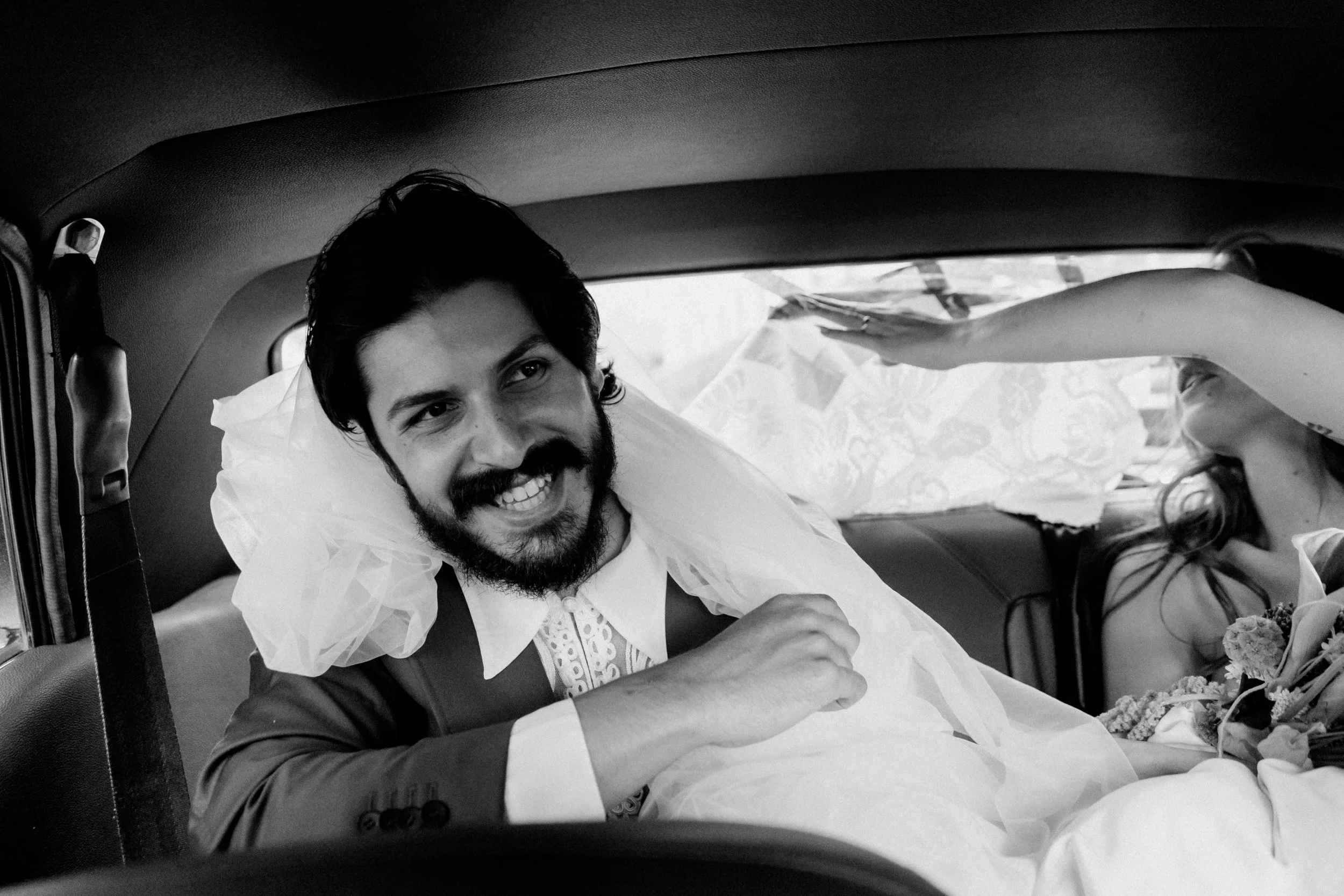 A man with dark hair, a beard, and wearing formal clothing, smiling and reclining inside a car, with a woman in the background reaching towards him, in black and white.