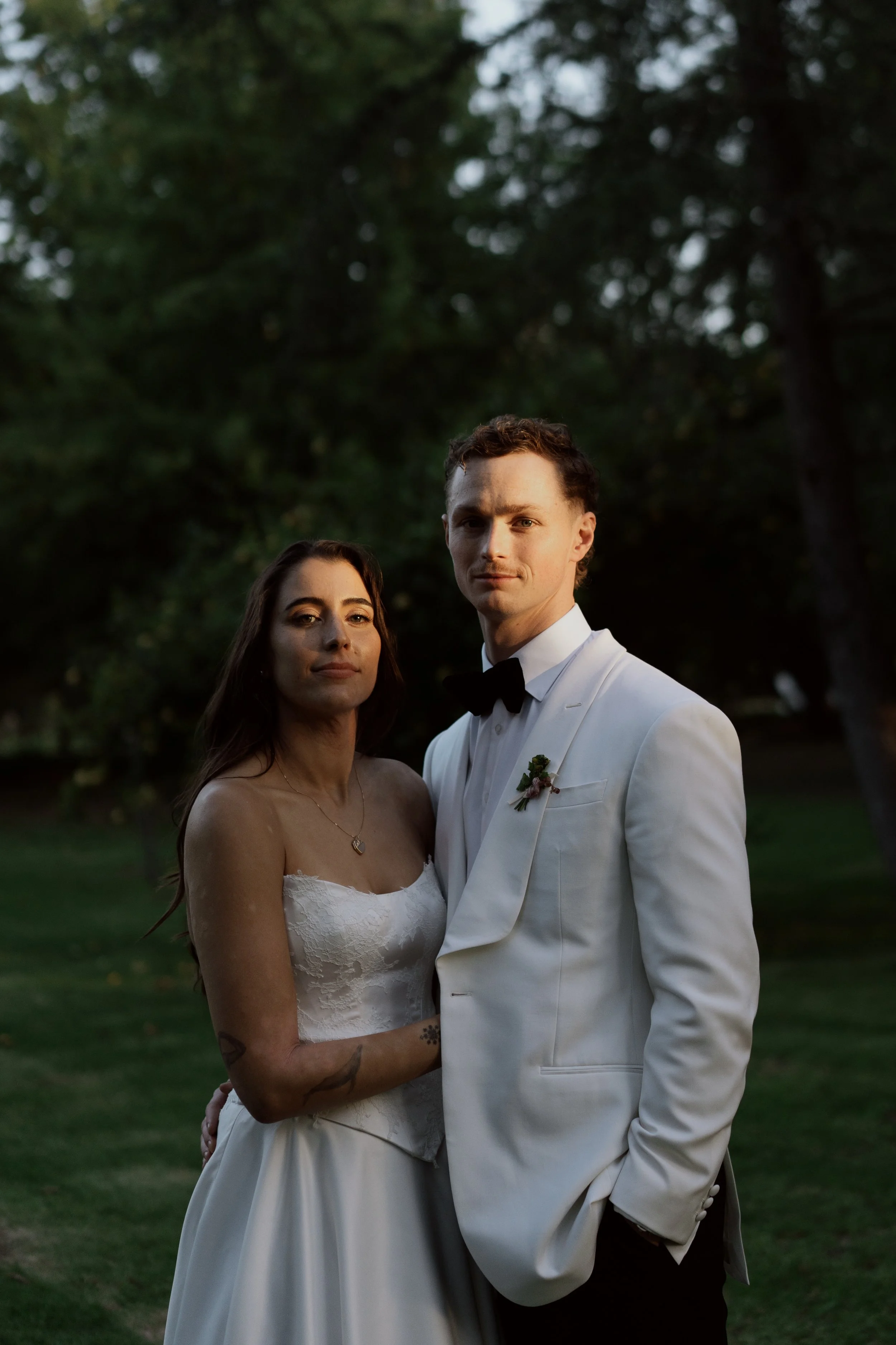 A couple in wedding attire standing outdoors during sunset, with trees in the background.