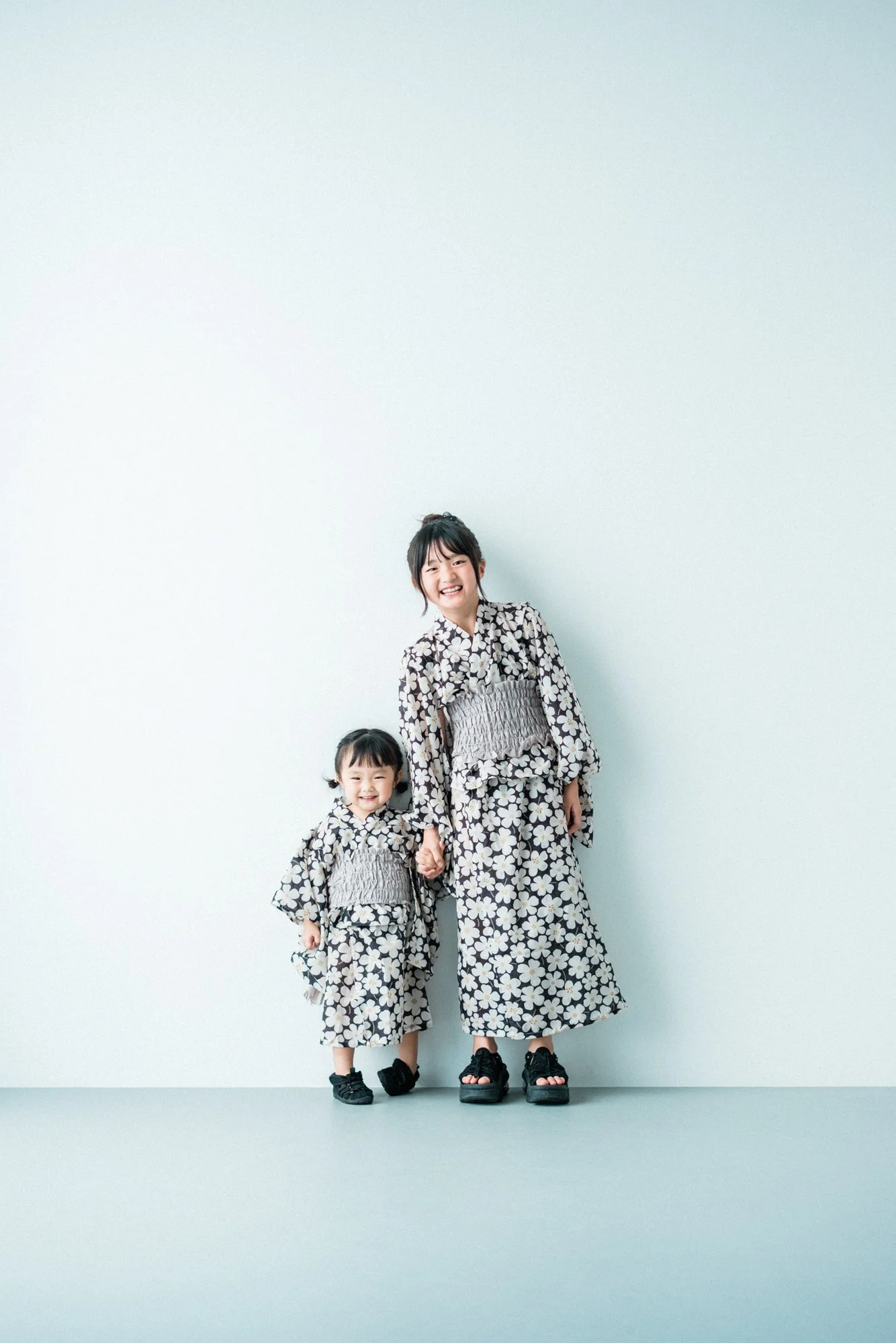 花柄の着物を着た少女と女性が手をつないで笑顔で立っている。背景は白い壁。