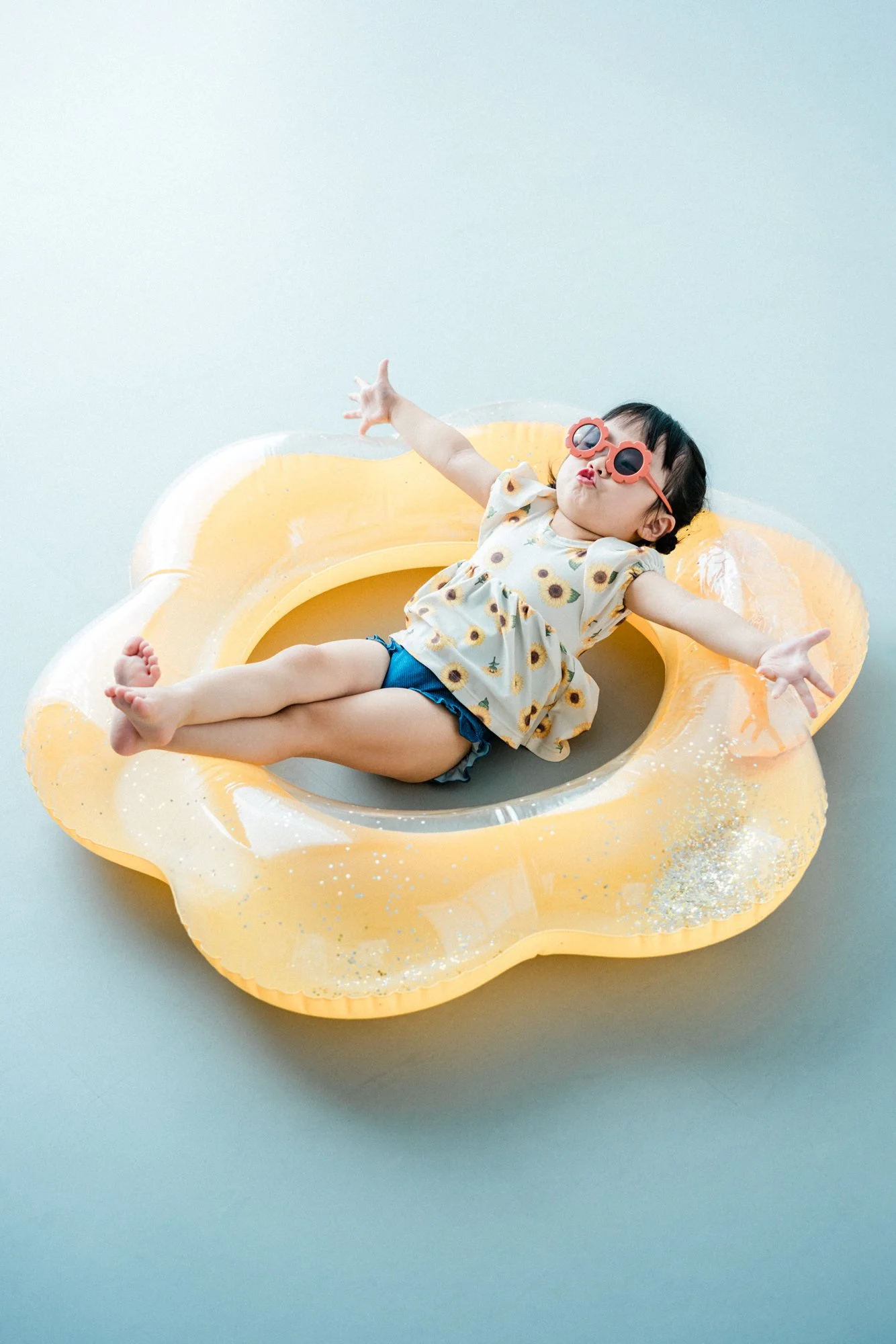 ビーチボールの形をしたプールの縁に座る子供。サングラスをかけ、花柄のトップスと青の短パンを着ている。