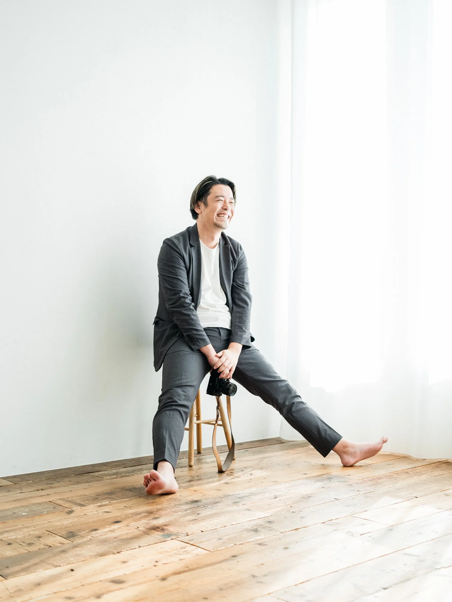 A man in a gray suit and white shirt sitting on a wooden stool, laughing, with one foot on the floor and the other extended outward, holding a camera in his right hand, in a bright room with white walls and wood flooring.