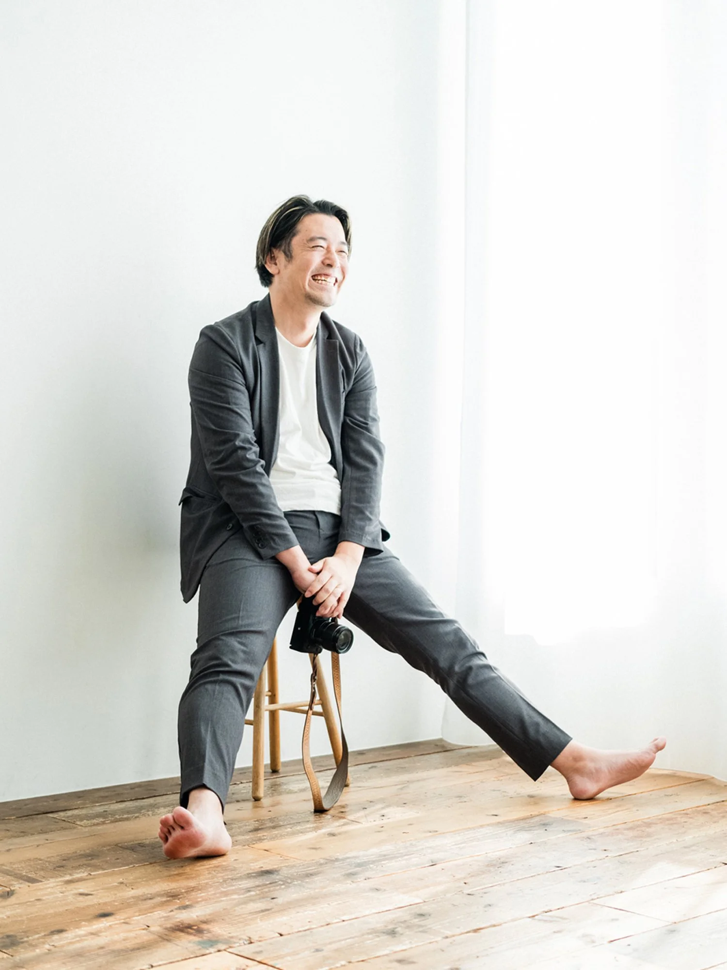 A man in a gray suit and white shirt sitting on a wooden stool, laughing, with one foot on the floor and the other extended outward, holding a camera in his right hand, in a bright room with white walls and wood flooring.