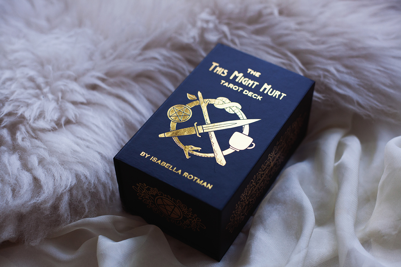  Image shows the front of This Might Hurt Tarot box on a background of white cloth and fur. Box is black with title and logo in gold foil. First printing. 