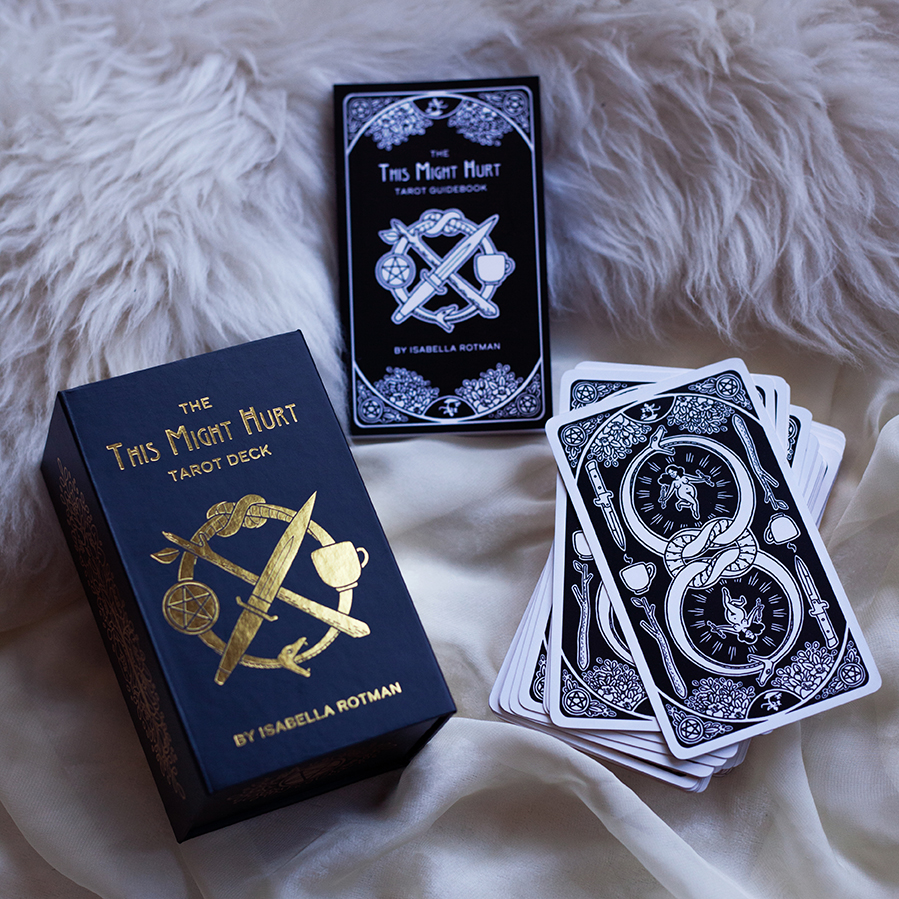  Image shows This Might Hurt Tarot box, guidebook, and card deck with backs facing the viewer. The guidebook is title “Title Might Hurt Tarot Guidebook” and decorated with florals and the logo. The card backs are decorated with a double ouroboros, fl