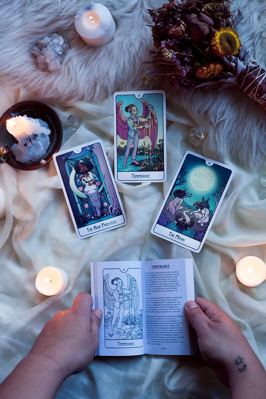 Image shows three cards surrounded by crystals and candles, with the guidebook being held open by hands beneath them. The guidebook is open to the card description for Temperance.