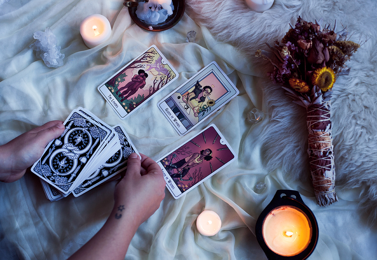  Image shows four cards surrounded by dried flowers add candles, with hands pulling another card from a deck of card backs. Cards are The Lovers, the Queen of Wands, and 7 of Wands. First printing. 