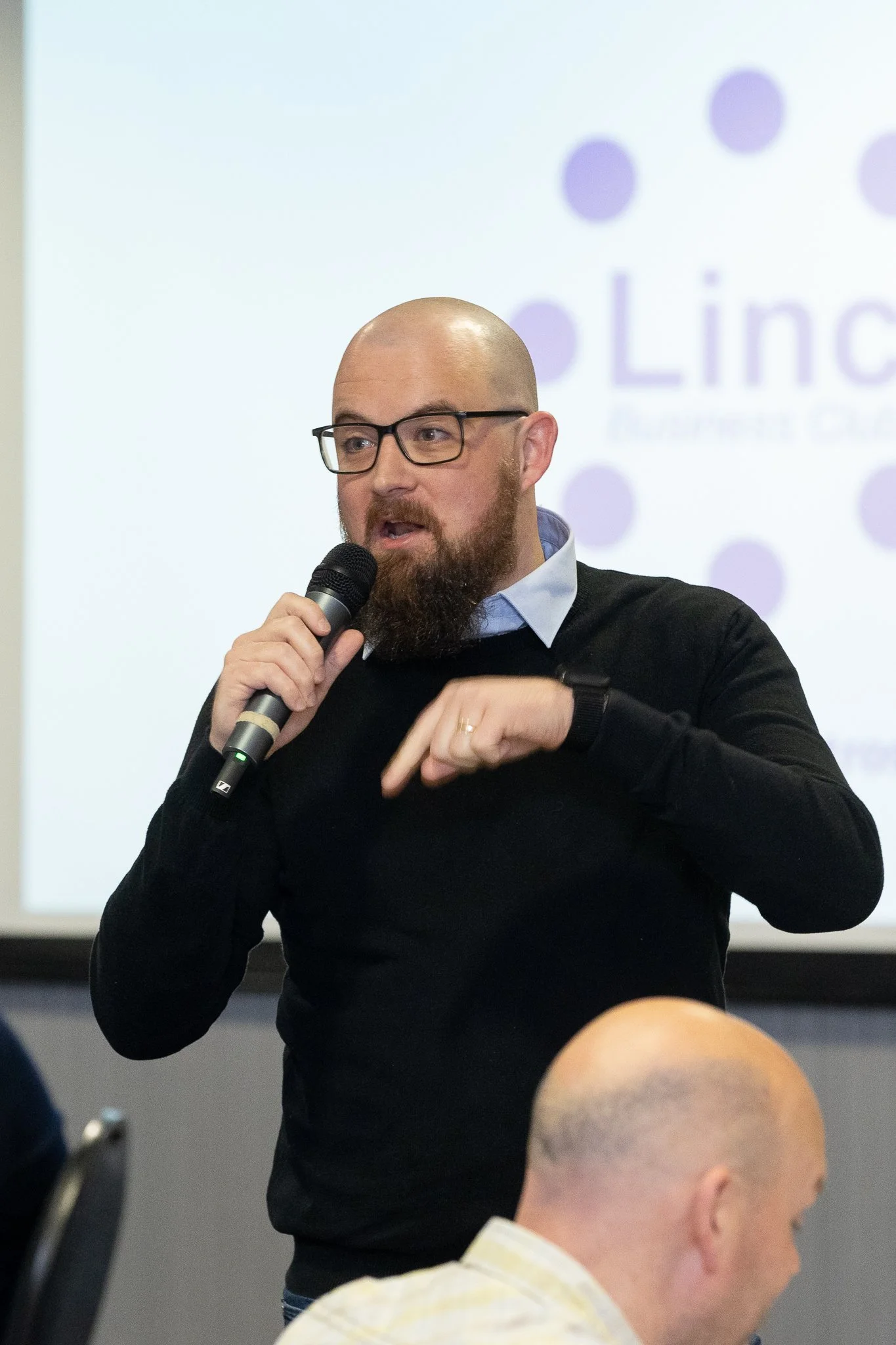 A man – Adam Driver, founder and MD at Authentic Comms Strategic Consultancy –  with glasses and a beard, wearing a black sweater and a light blue collared shirt, speaking into a microphone during a presentation or talk.