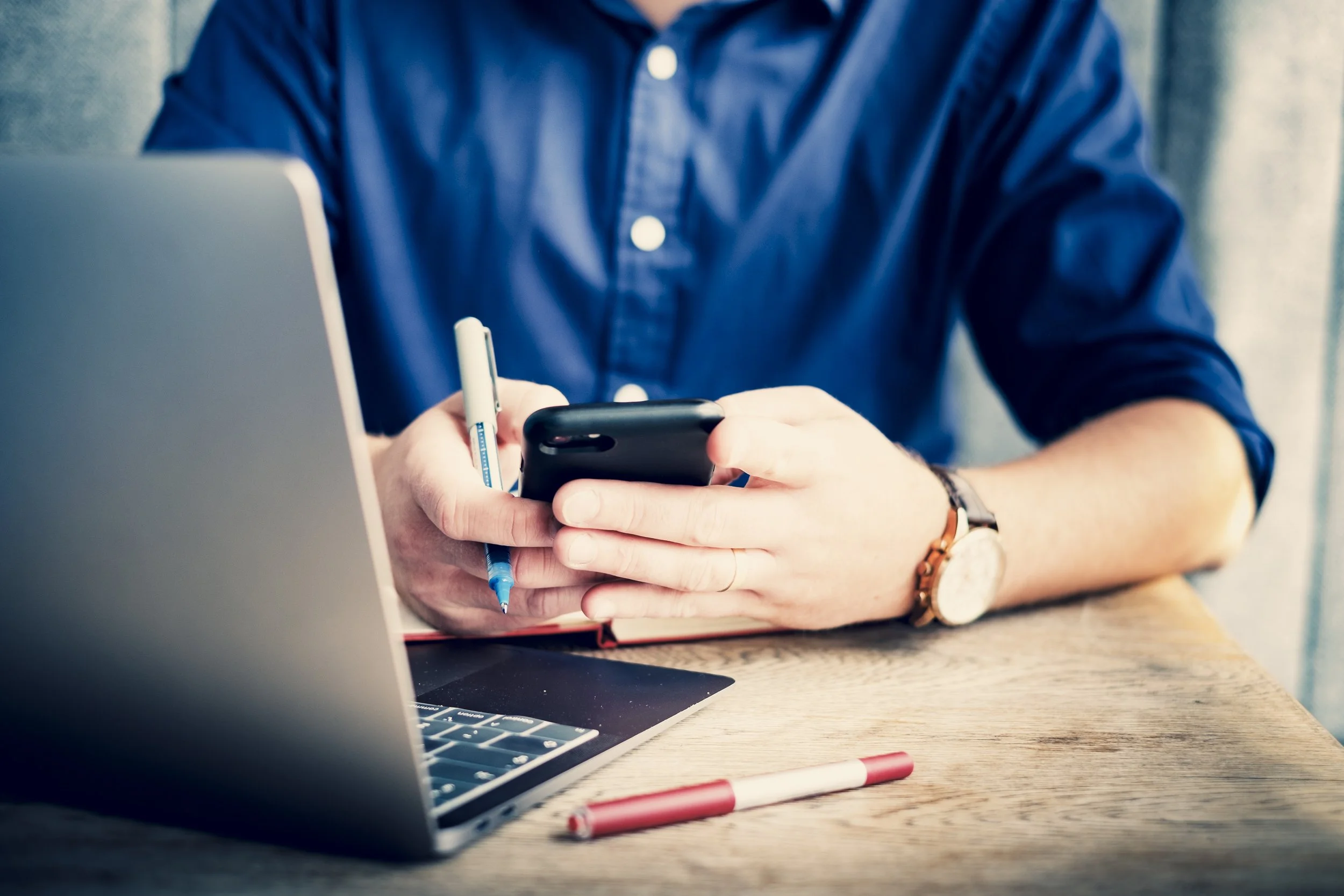 Marketing consultant in blue shirt holding a phone, with a laptop and red pens on wooden table.